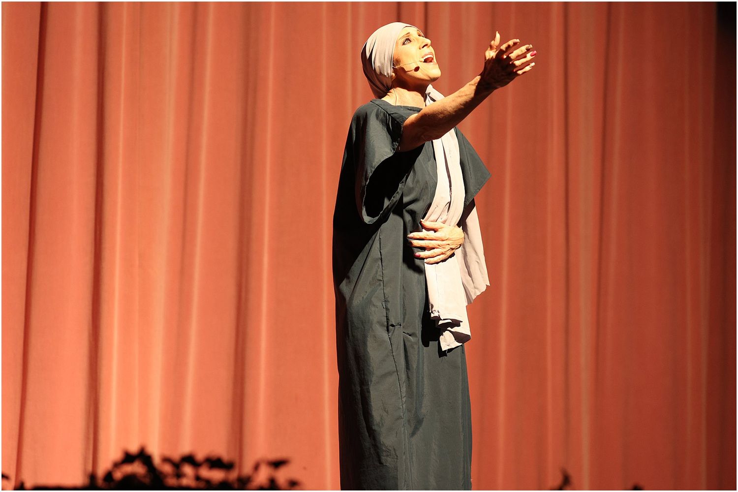 Stage performance photo showing animated speaker in gray outfit against red curtain backdrop.