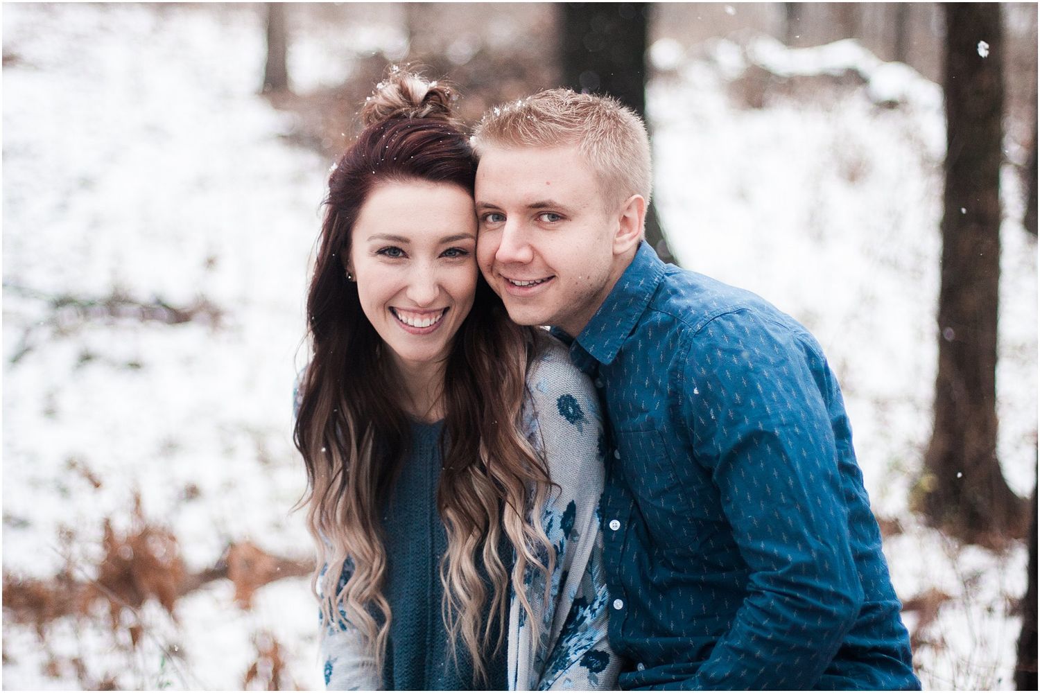 A joyful winter portrait of a couple wearing coordinating blue outfits in a snowy forest setting.