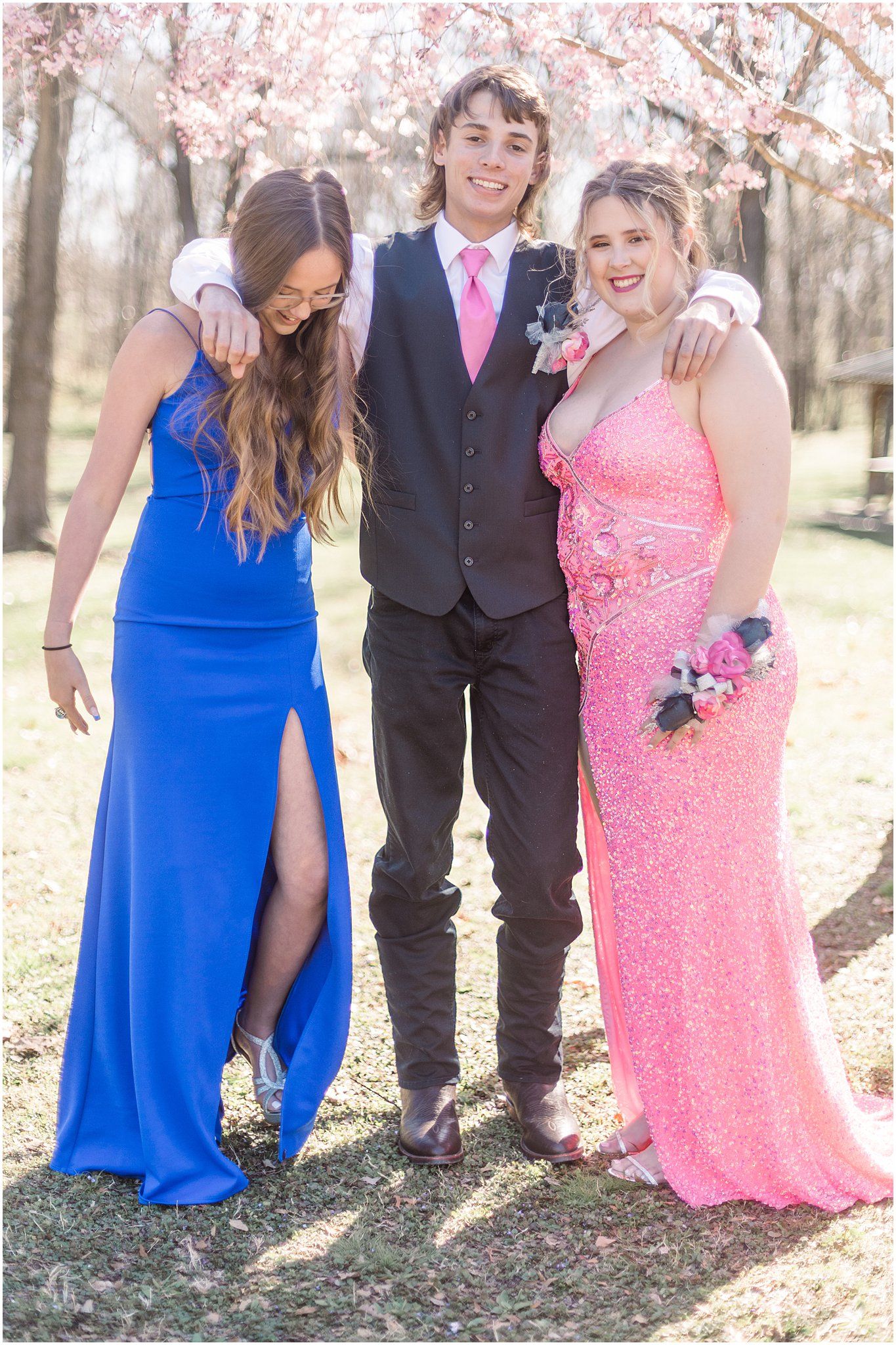Three people in prom attire smile together under cherry blossoms wearing blue, black, and pink formal wear.