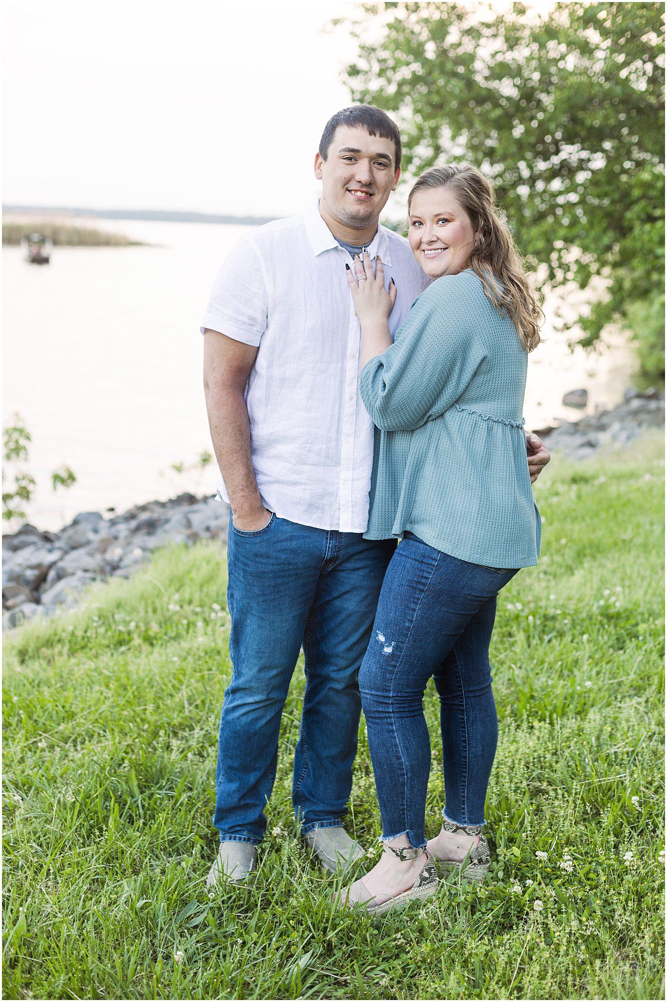 Casual couple portrait by waterfront with rocky shoreline and grass in background.