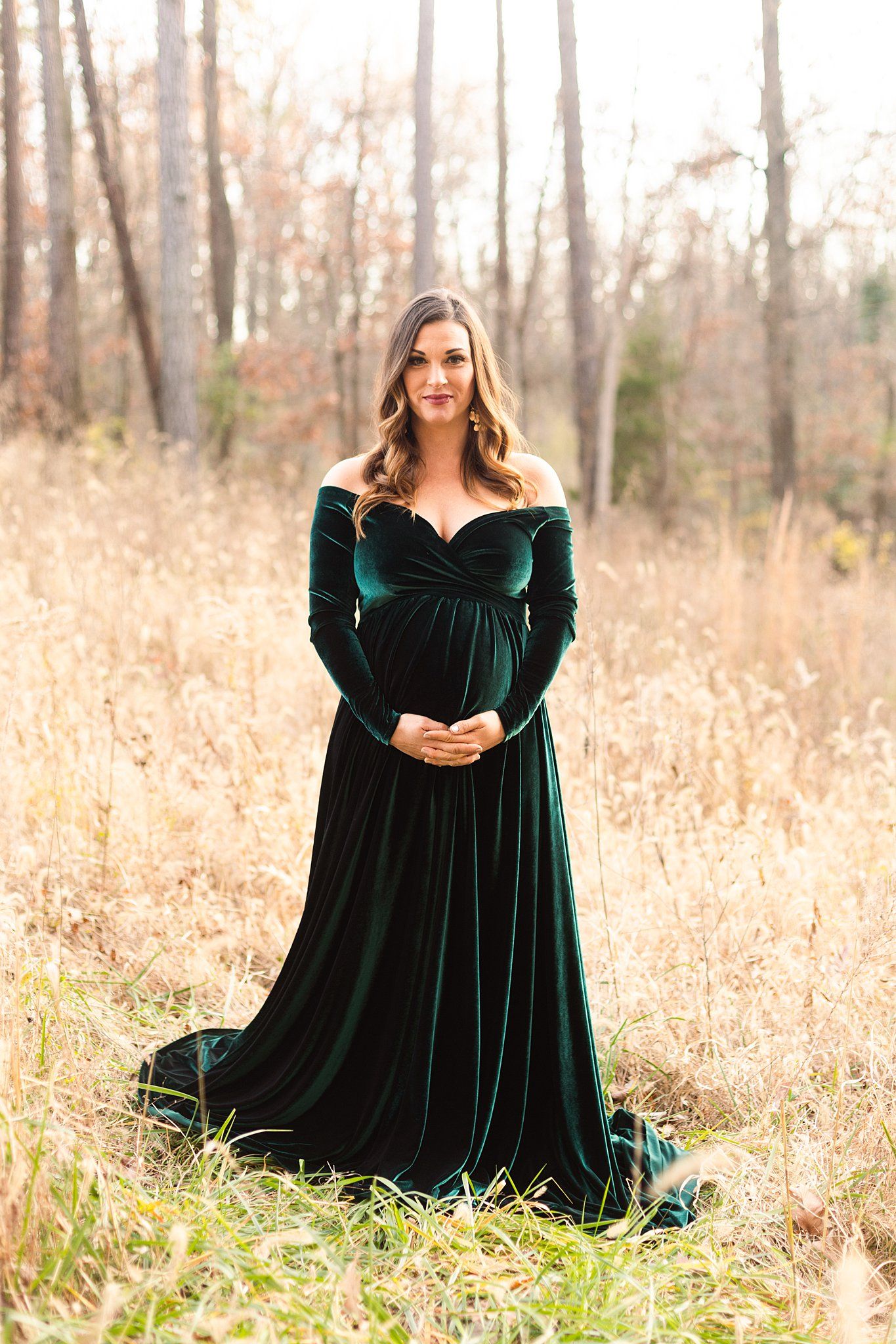 Beautiful emerald velvet maternity gown worn in an autumn field with soft sunlight filtering through bare trees.