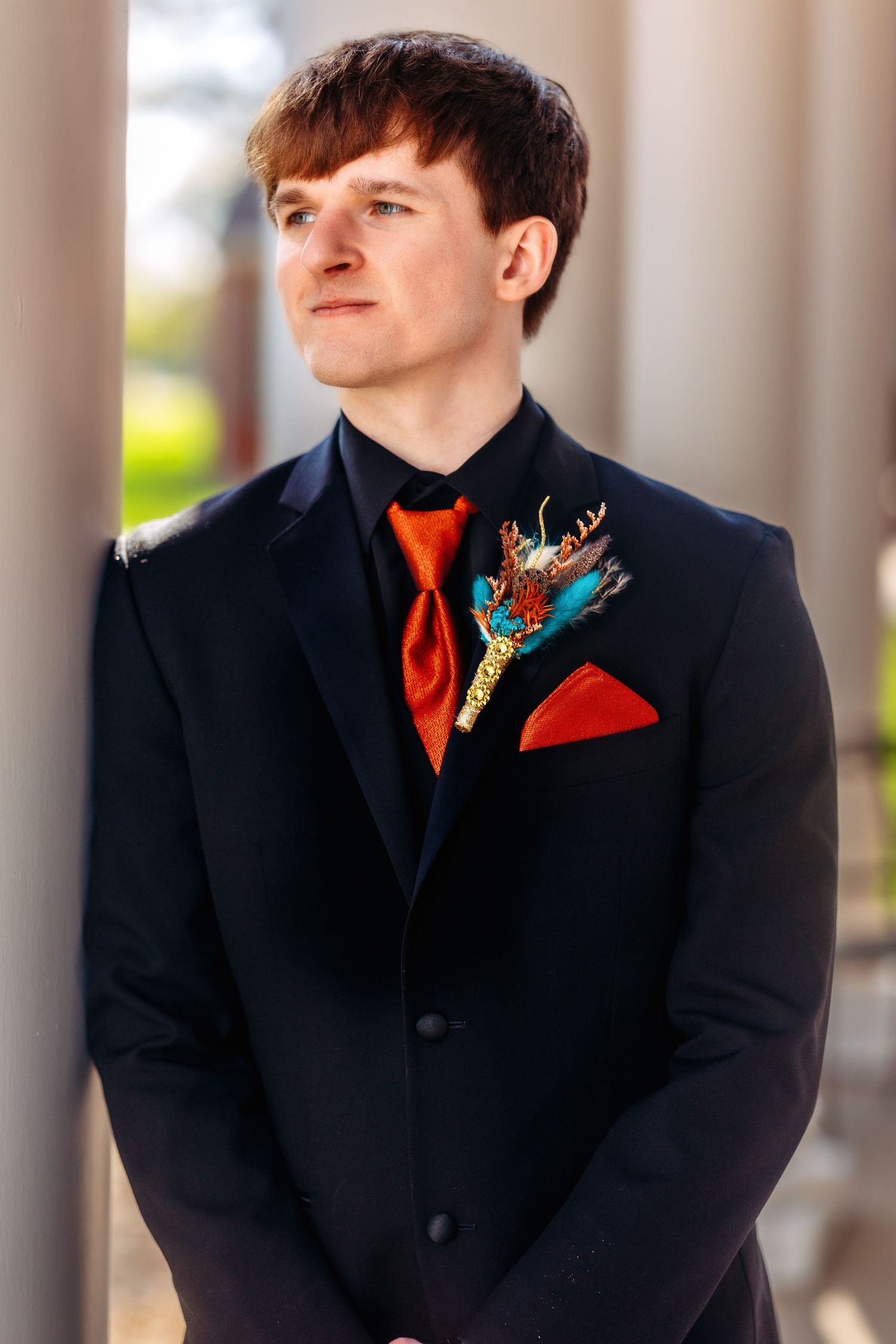 A person in a black suit with an orange tie and floral boutonniere poses against white columns in a formal setting.