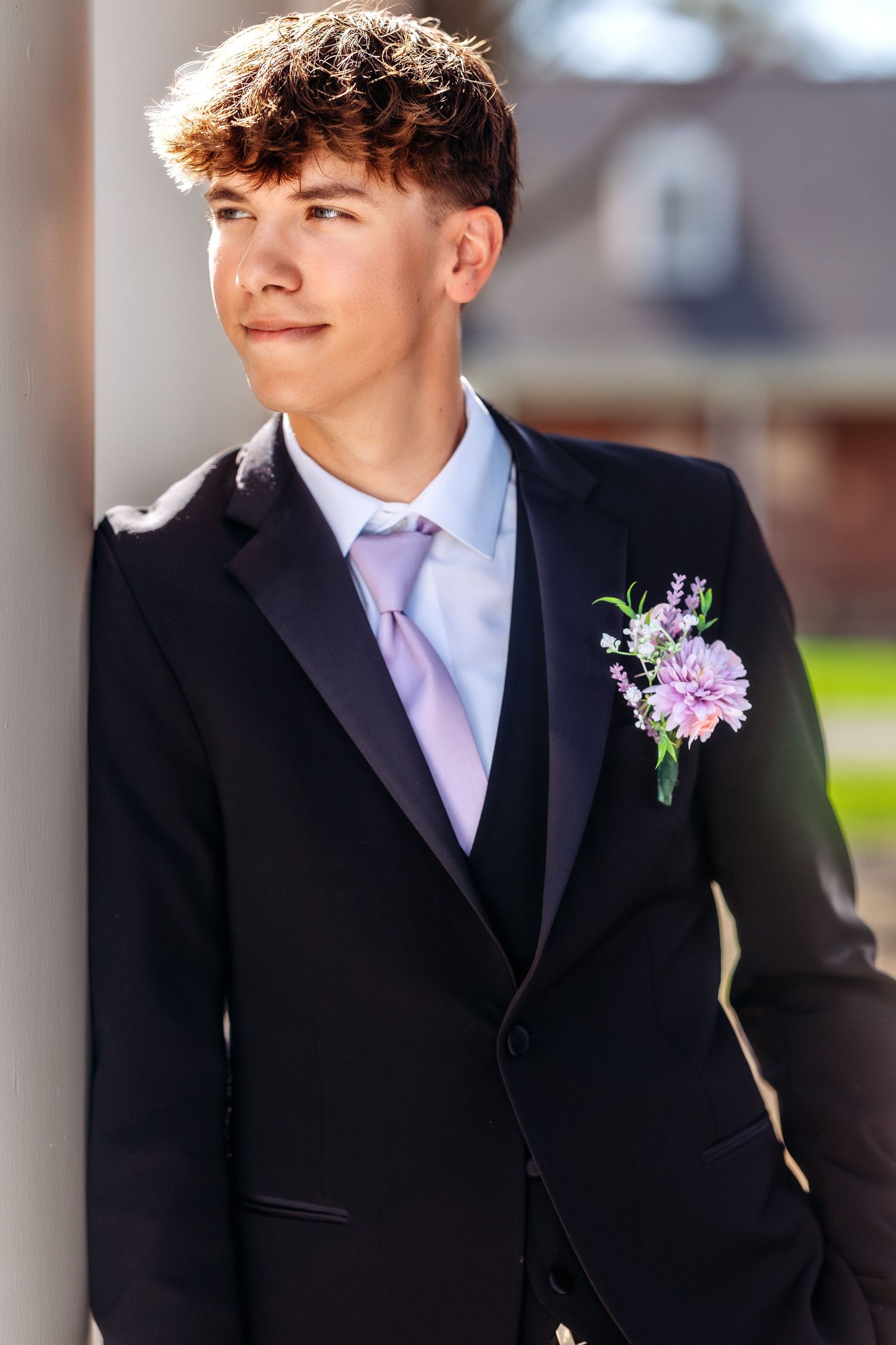 A person in a black tuxedo with a pink and purple floral boutonniere and light pink tie smiles while standing outdoors.