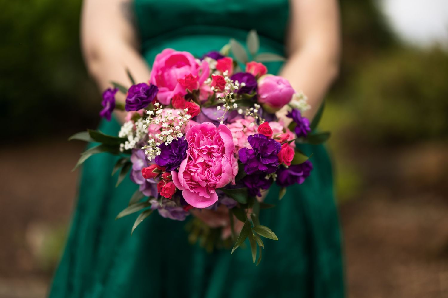 Woman in green dress holding vibrant pink and purple peony wedding bouquet with greenery accents.