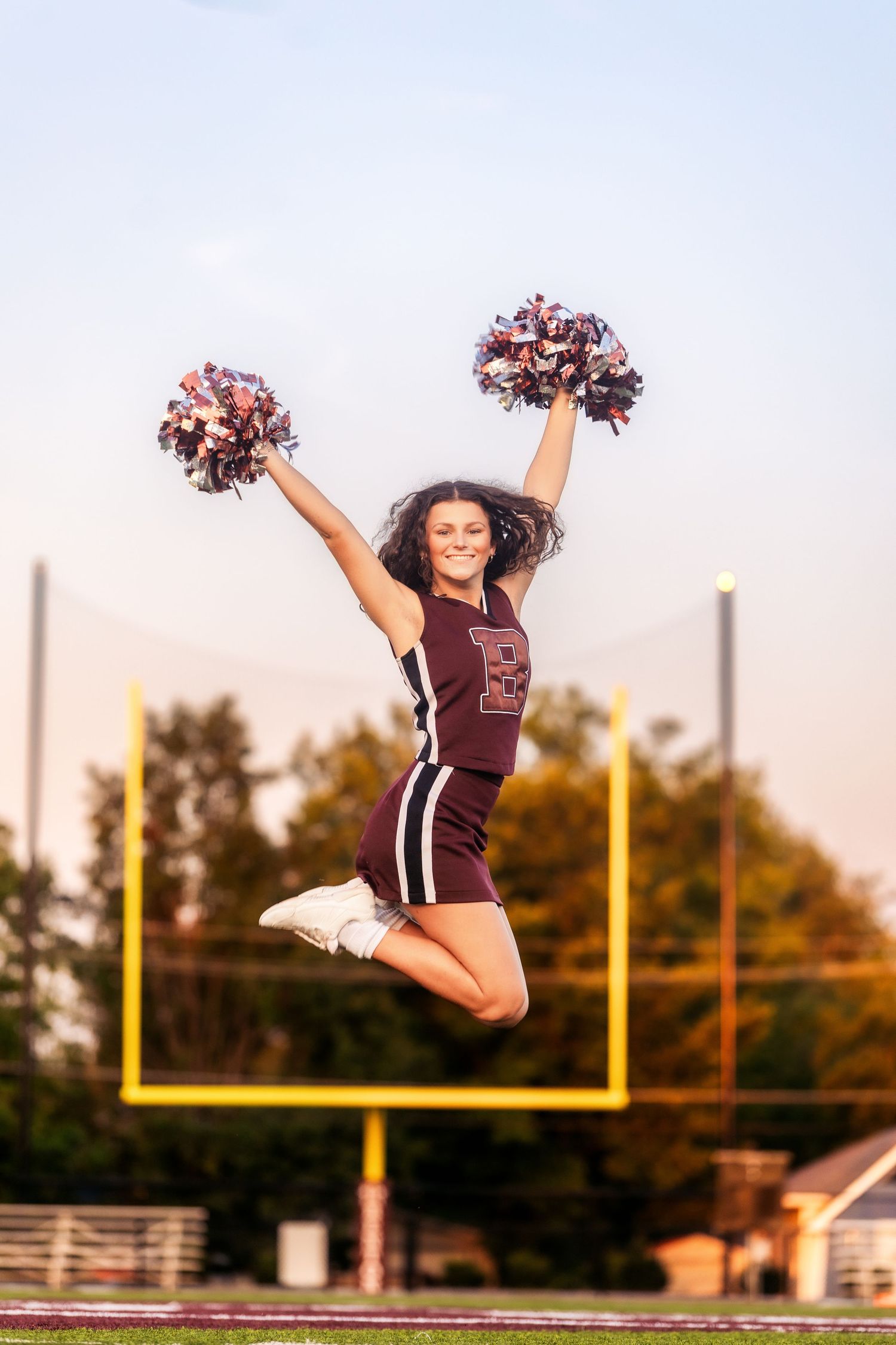 Cheerleader in maroon uniform jumping with pom-poms raised at football field during golden hour.