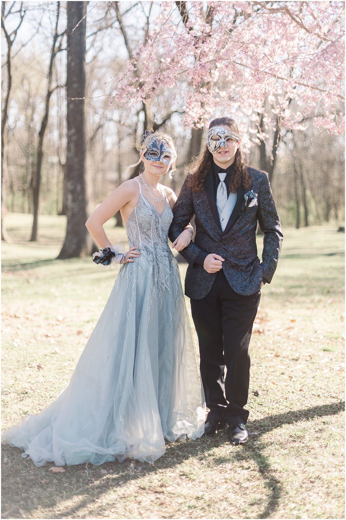 Couple in elegant formalwear and decorative masks stand together among cherry blossoms.