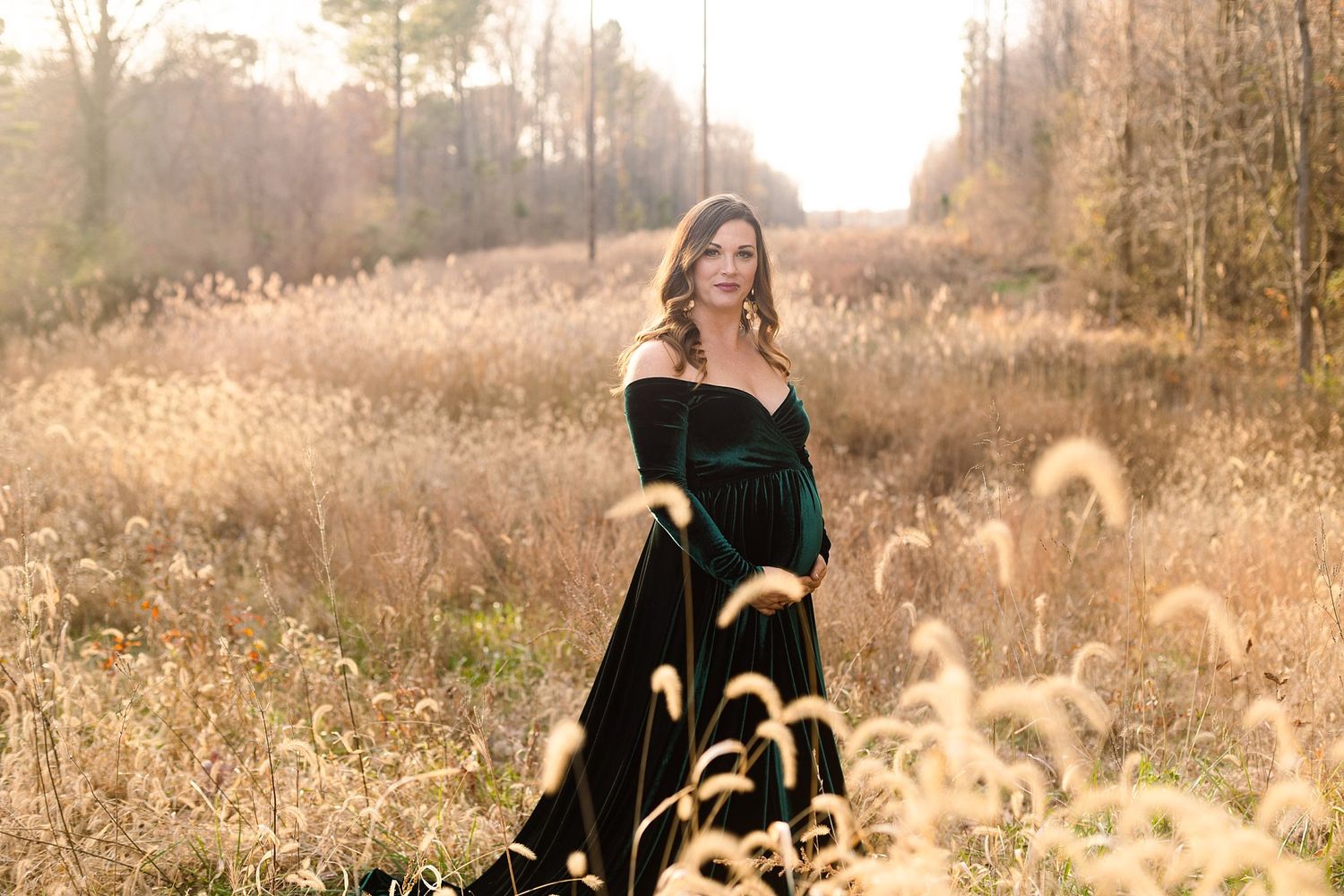 Woman in off-shoulder emerald velvet gown poses in sunlit autumn field with tall grass.