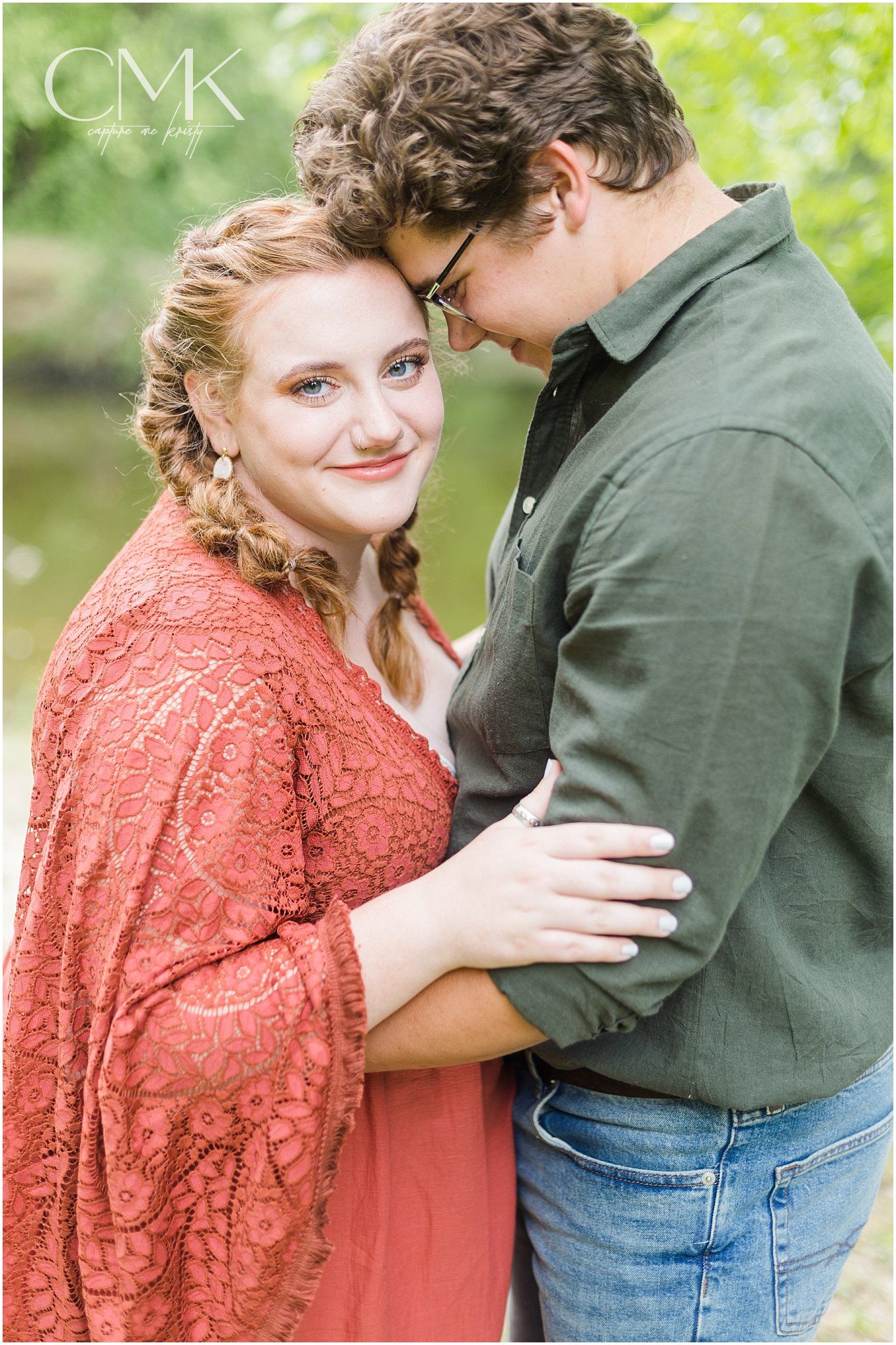 A romantic couple poses in an intimate embrace wearing casual attire against lush green foliage.
