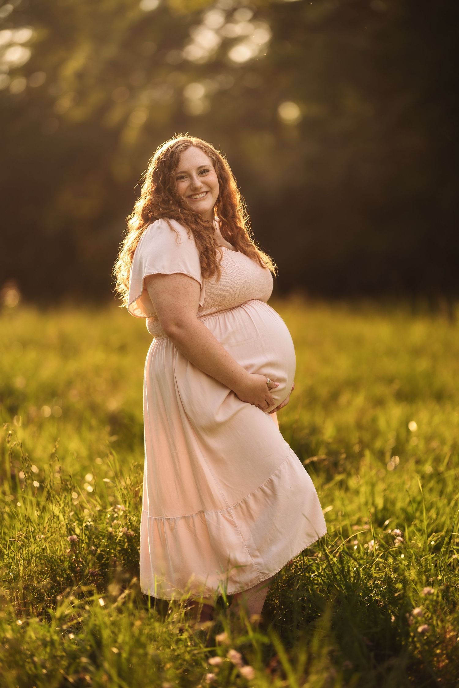 A maternity portrait in a pink dress during golden hour in a grassy field.