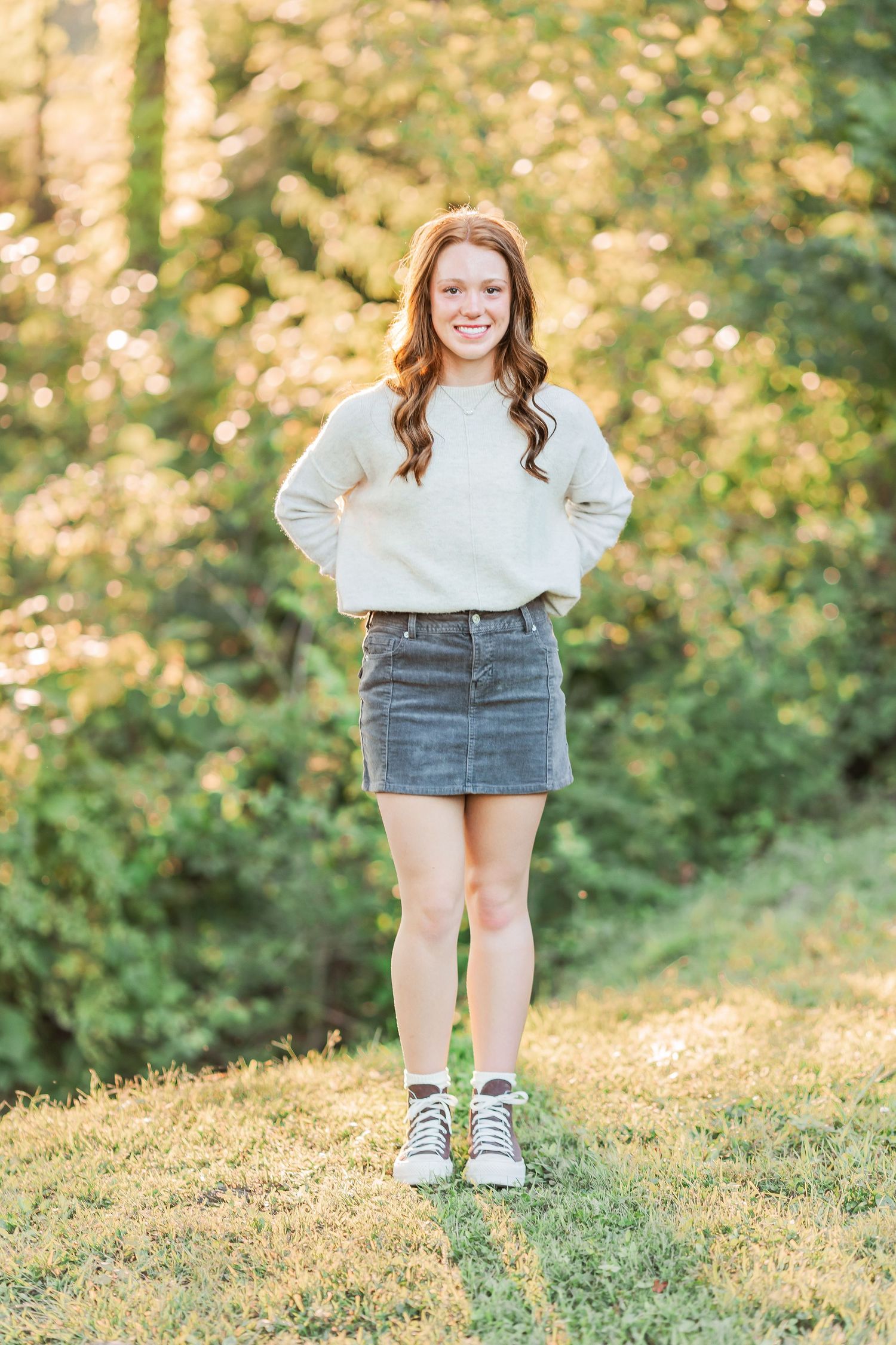Person in white sweater and denim skirt poses on grassy hill during golden hour.