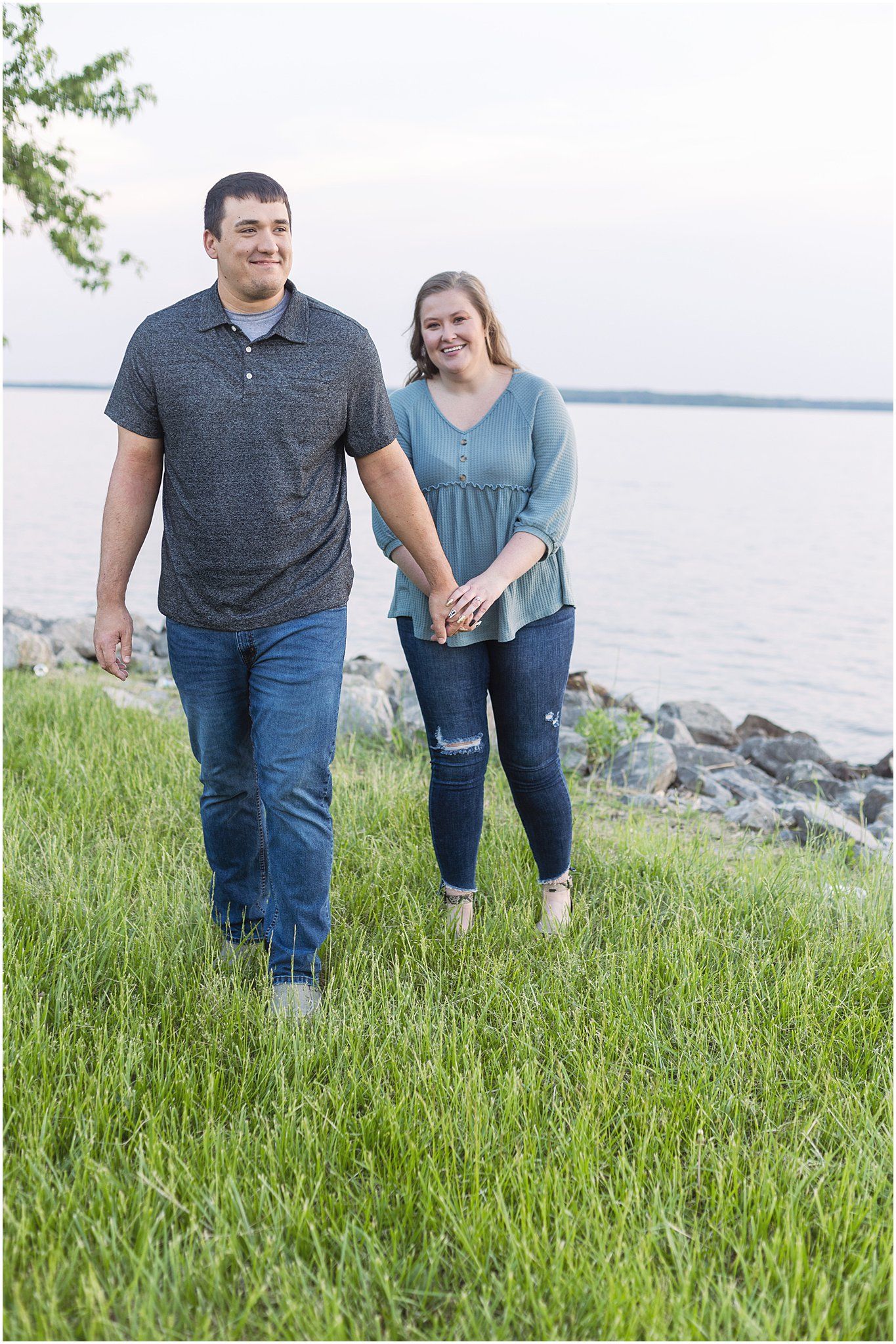 A couple walks hand in hand along a grassy waterfront path with a lake in the background.