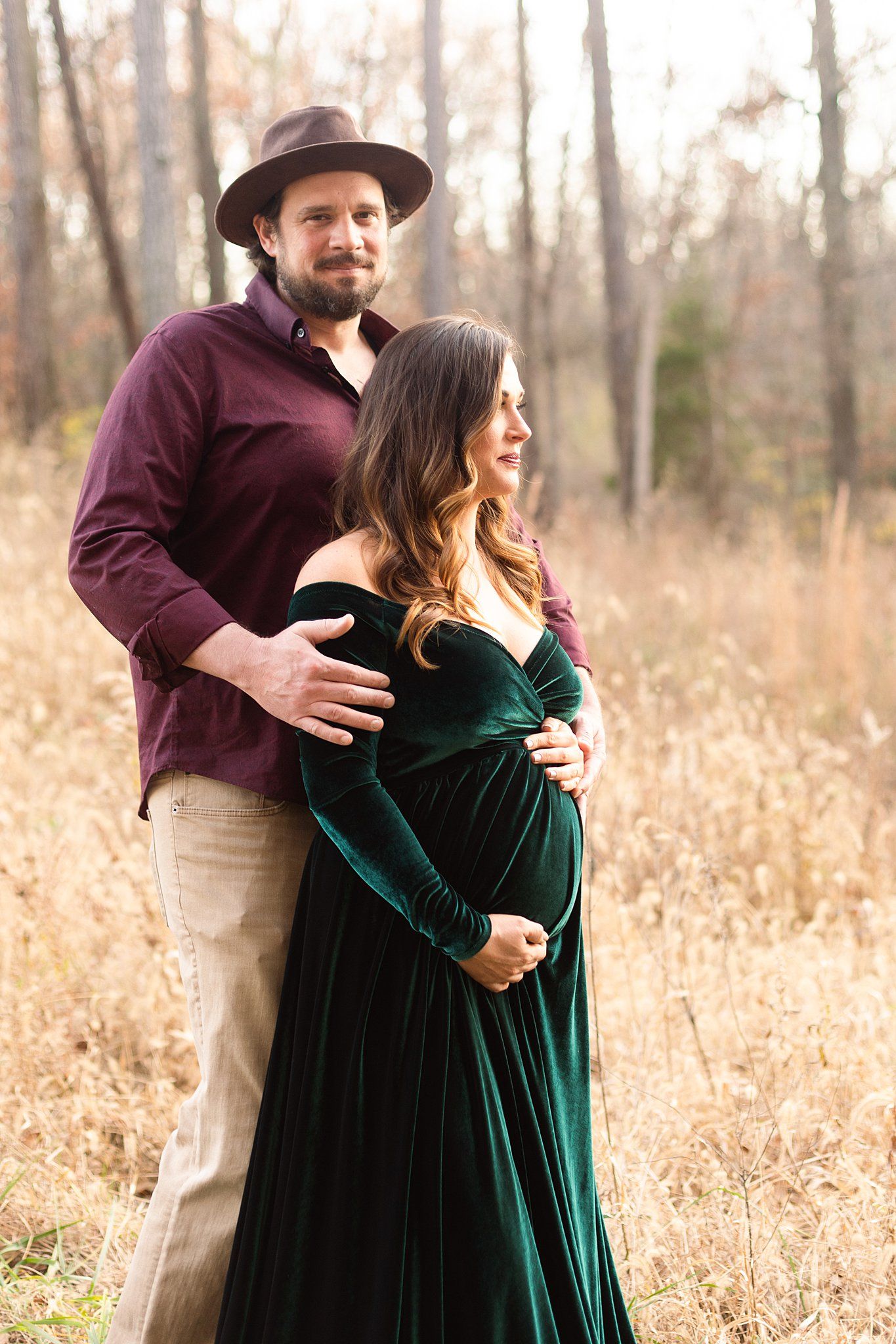 Couple embracing in autumn field wearing burgundy shirt and emerald velvet maternity gown.