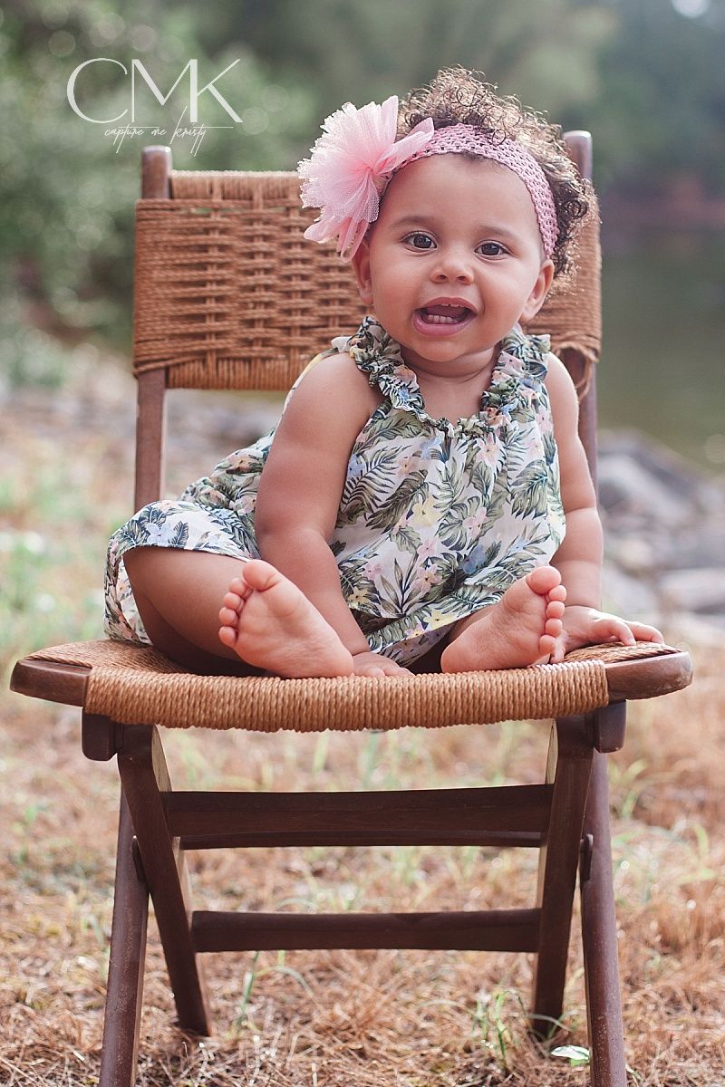 Baby sitting in wooden chair wearing floral dress and pink bow headband.