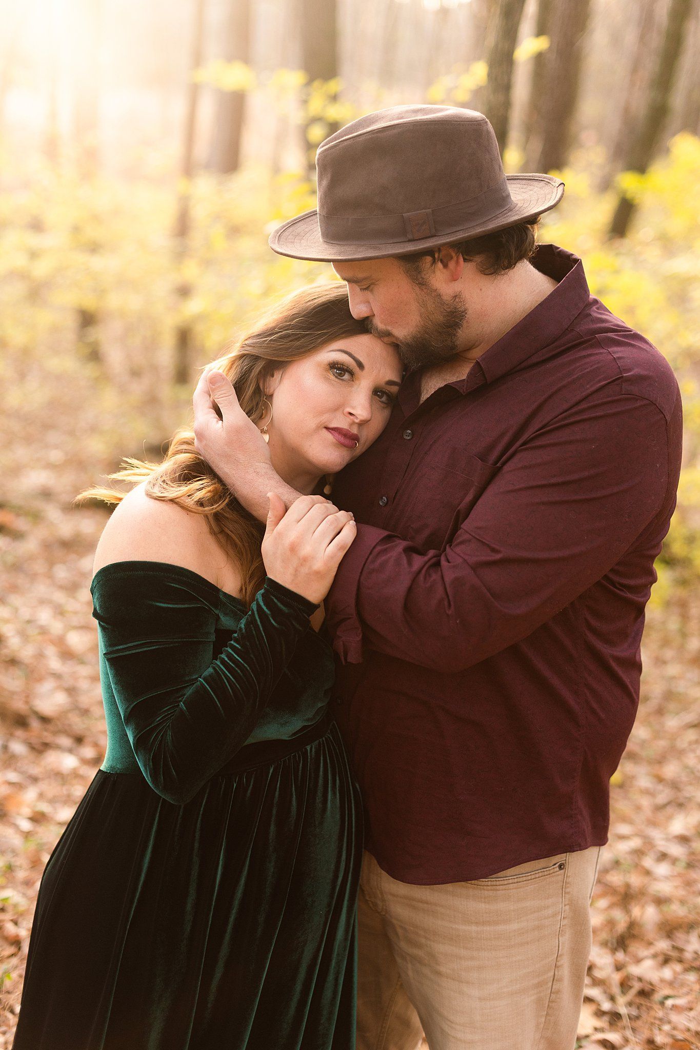 Tender embrace between couple in autumn woods wearing velvet dress and brown hat.