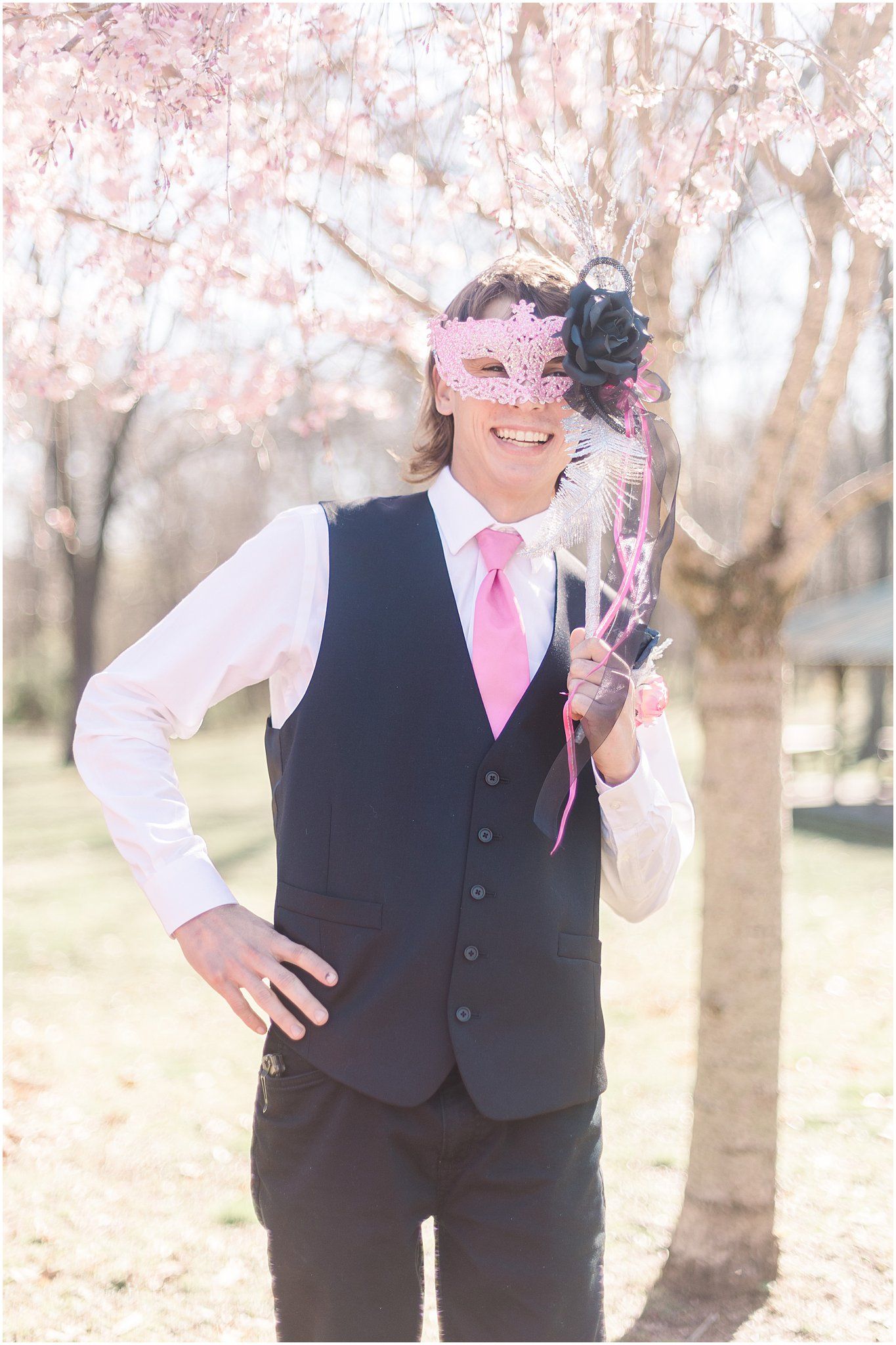 Person in formal vest and tie poses wearing decorative pink and black masquerade mask.