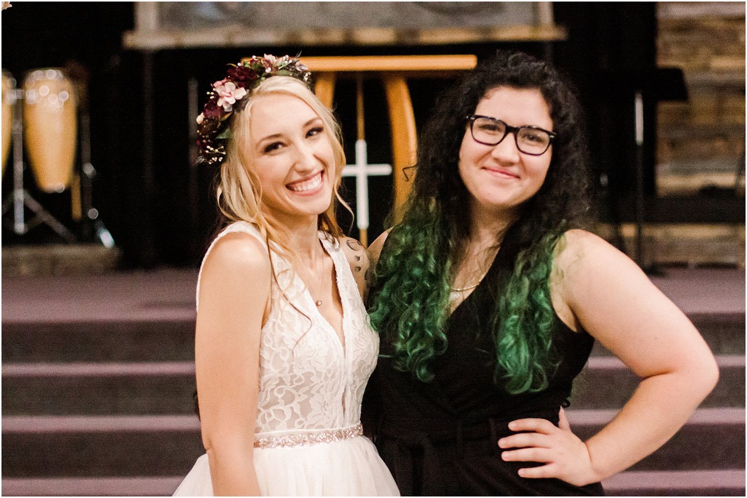 Two friends pose together at a rustic outdoor venue, one in a white wedding dress and one in a black dress.