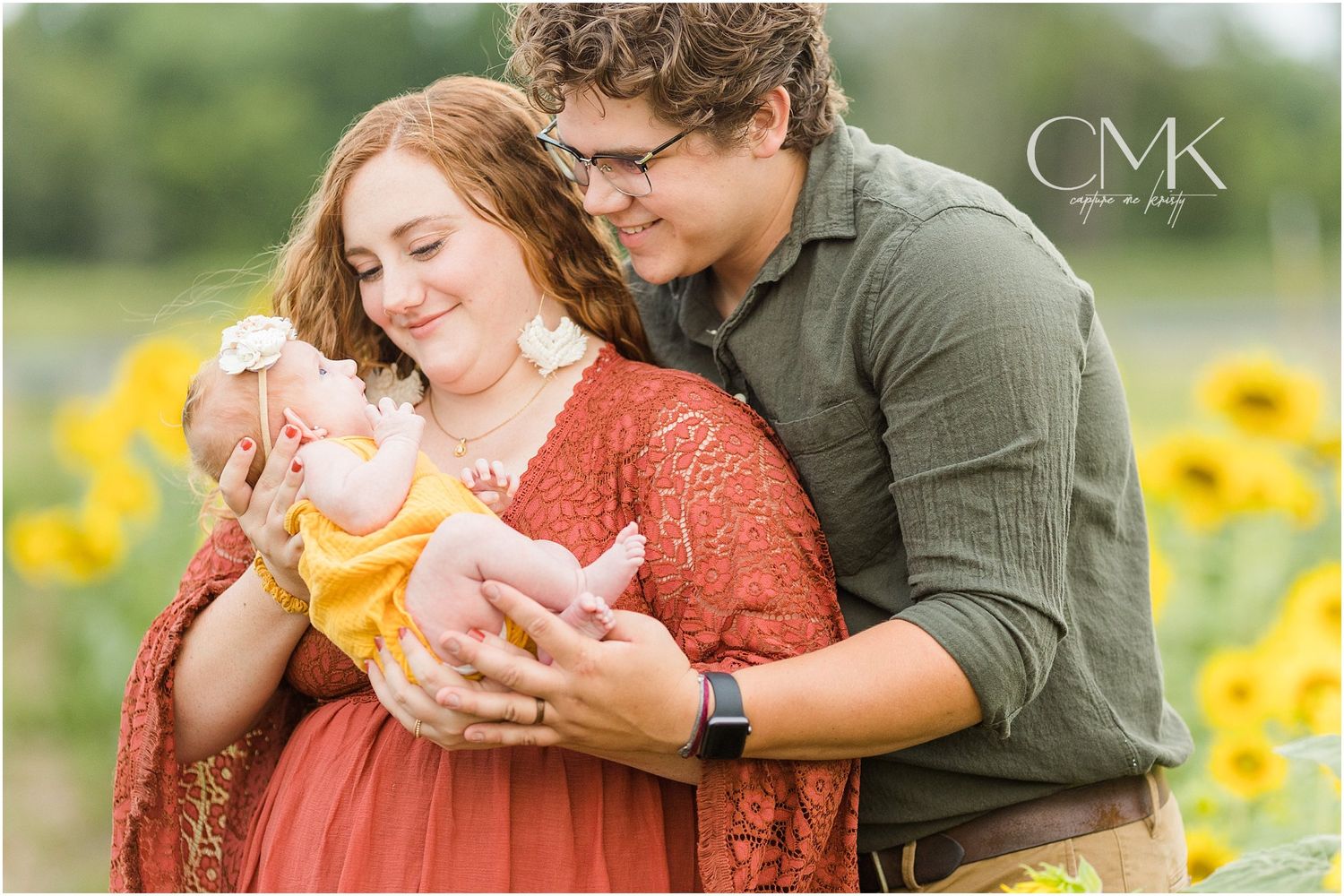 A family portrait in a sunflower field showing parents holding their newborn baby.
