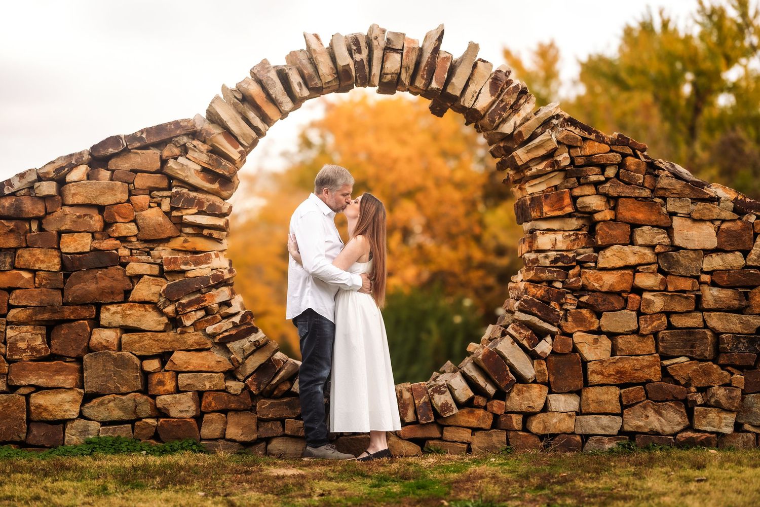 A couple in wedding attire poses romantically by a rustic stone wall during golden hour sunset.