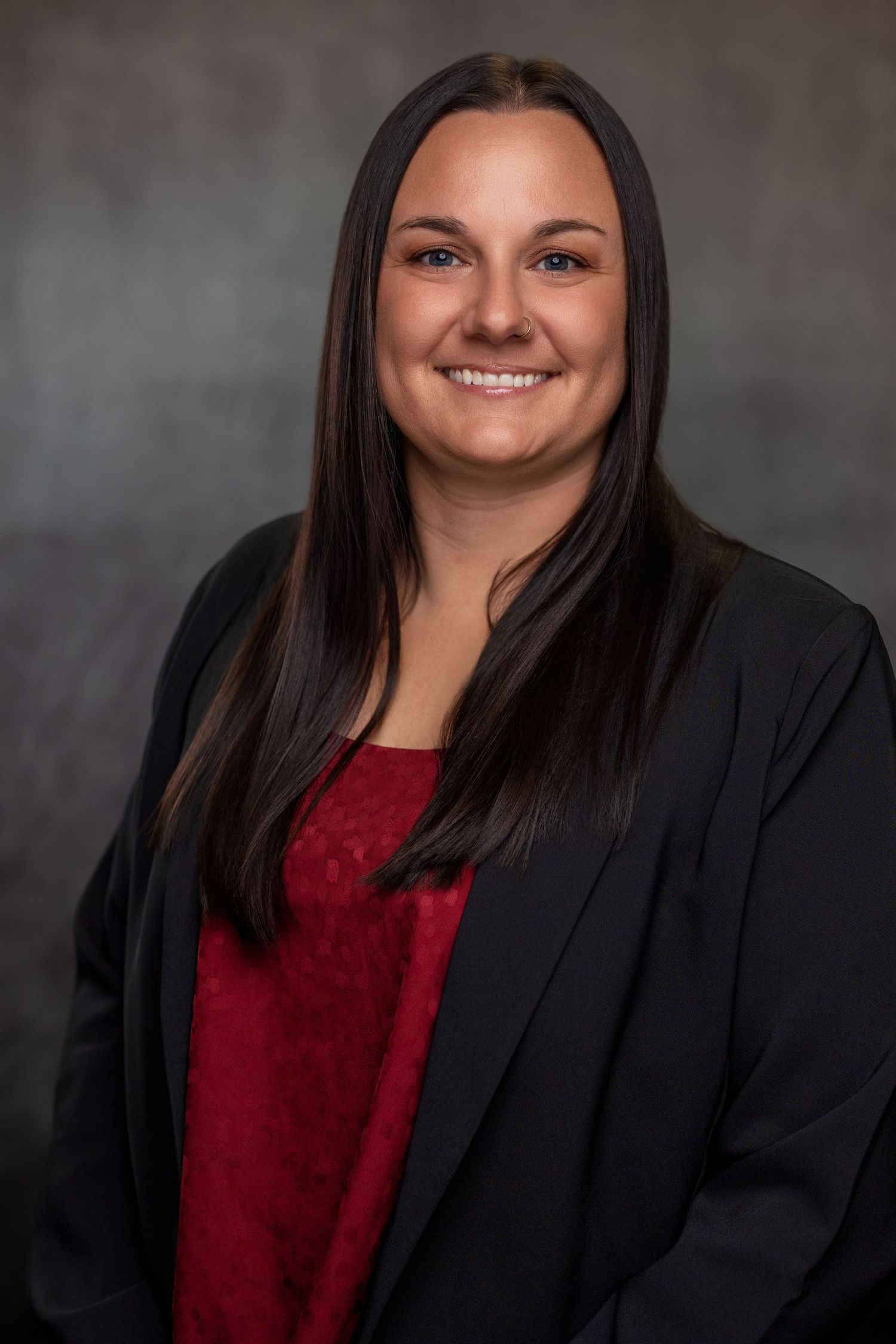 Professional headshot of woman in black blazer and red top against neutral background.