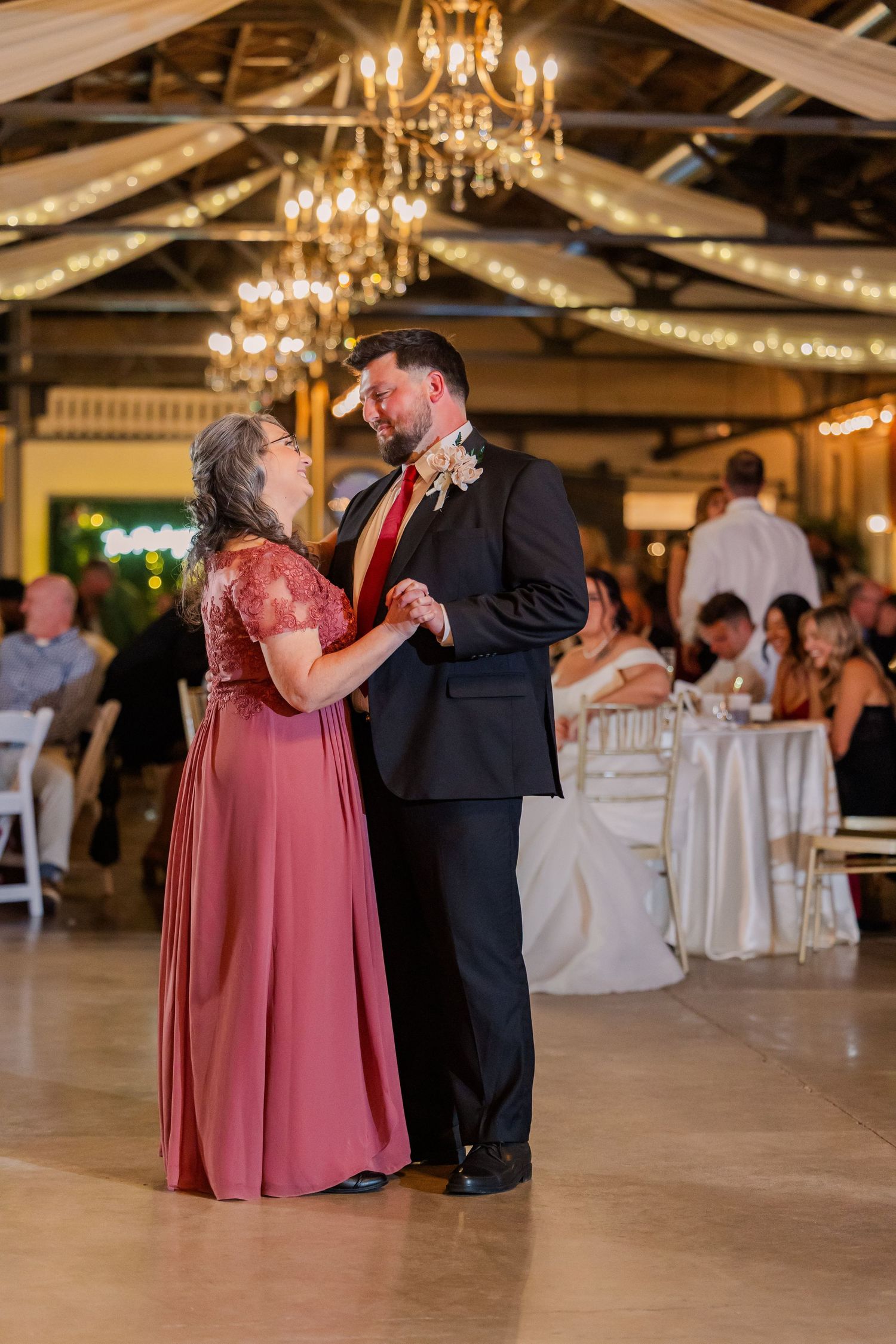 Couples dancing at a wedding reception under warm chandelier lighting in a rustic venue.