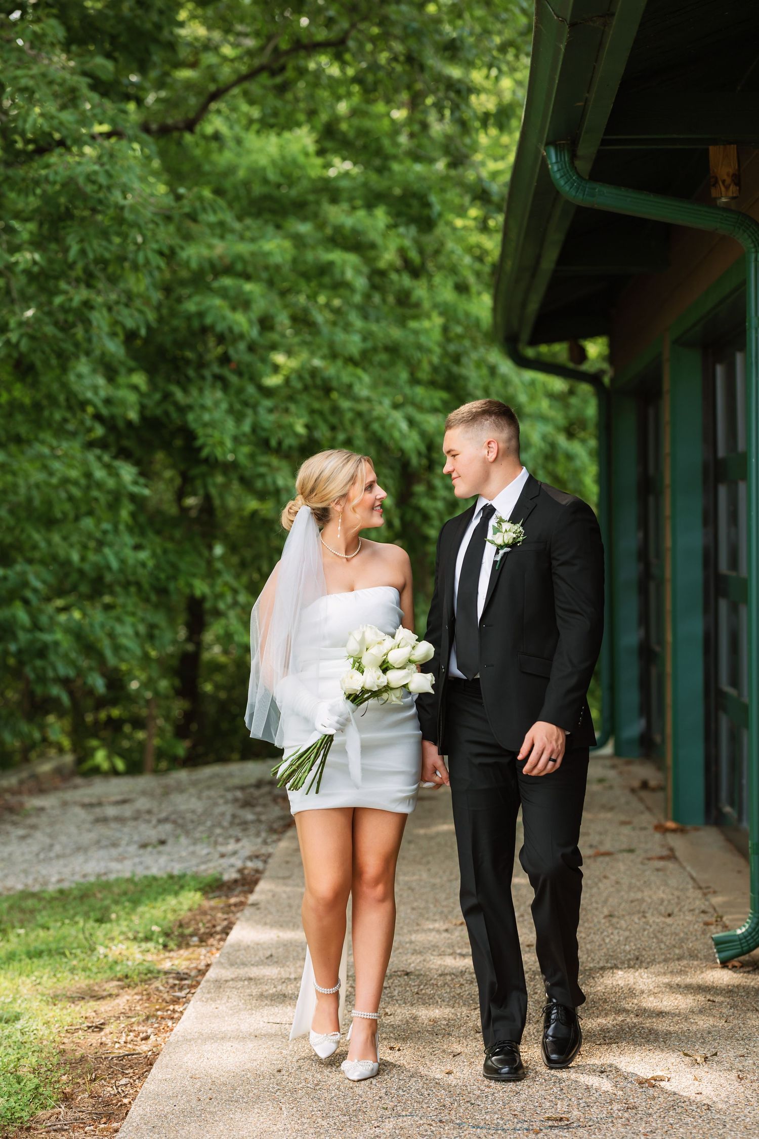 Wedding couple walking along covered pathway surrounded by lush green trees.