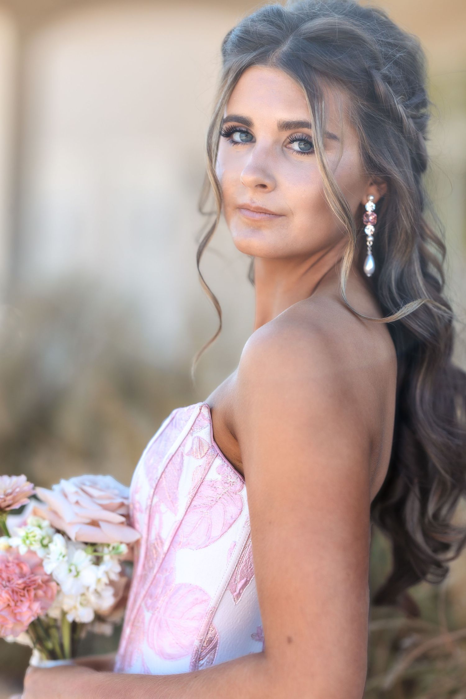 A formal portrait of someone in a pink gown with elegant upswept hair and dangling earrings holding a bouquet.