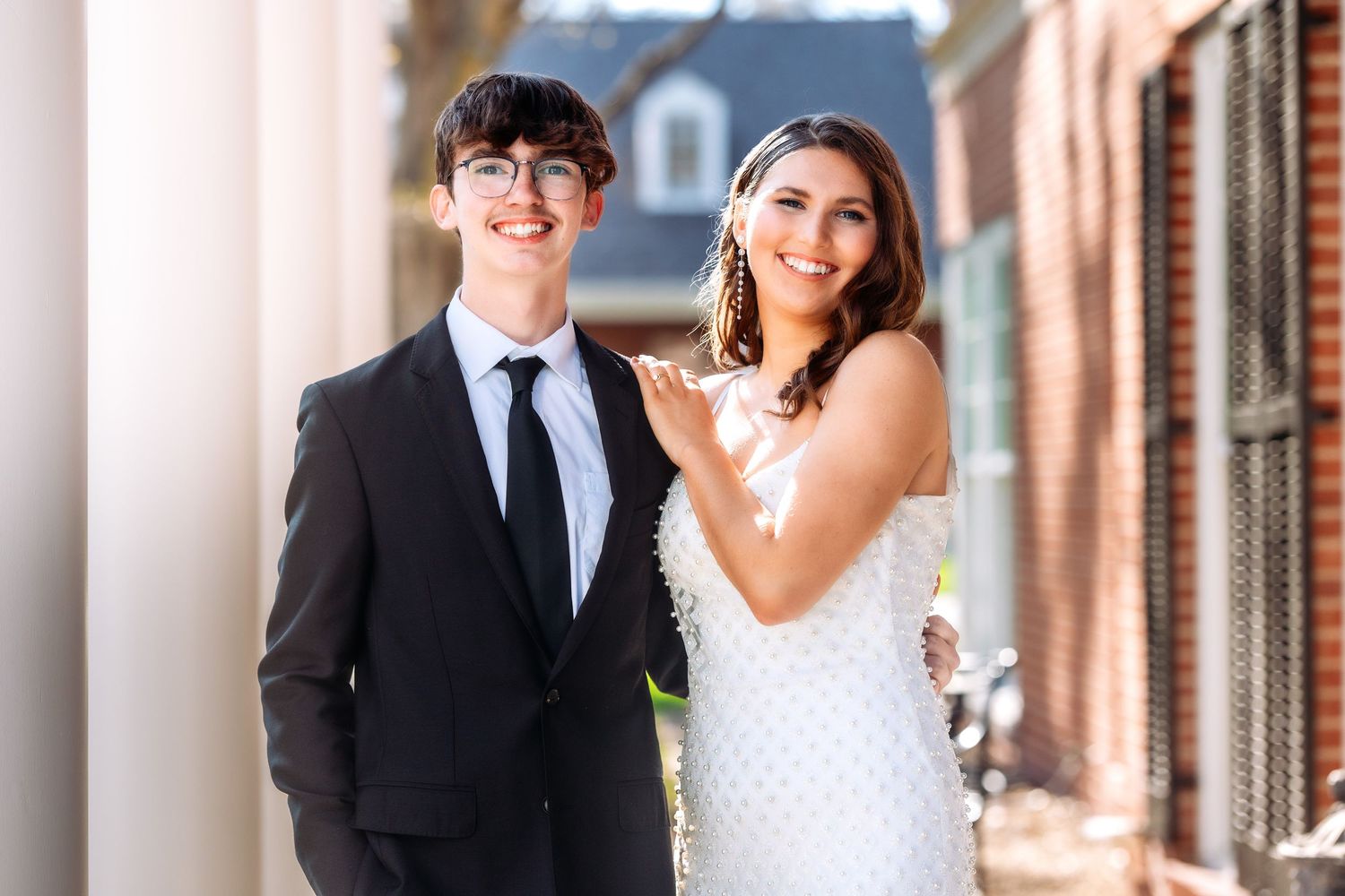A couple in formal wedding attire pose together on a brick sidewalk.