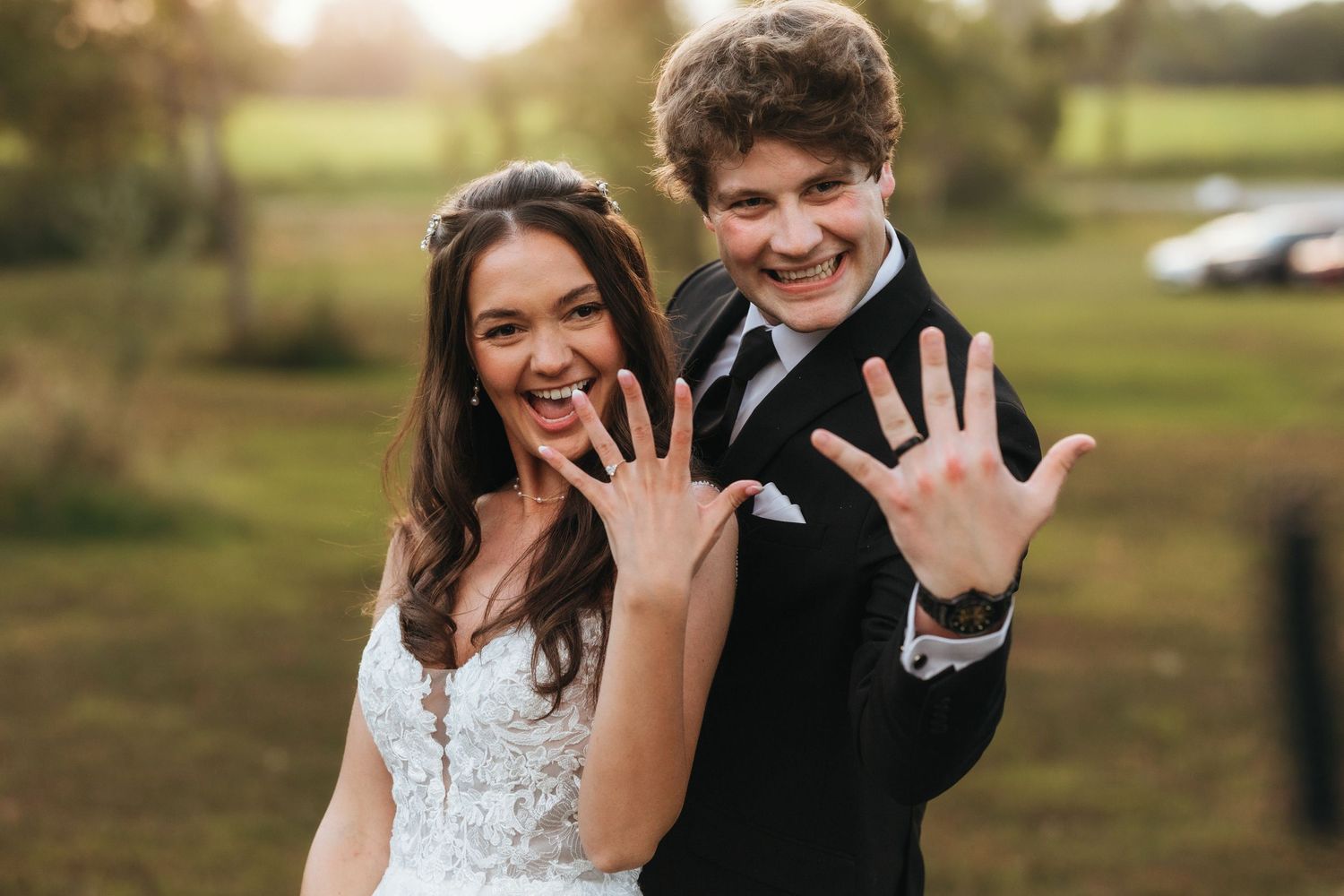 Newlyweds showing off their wedding rings with excited expressions in an outdoor setting.