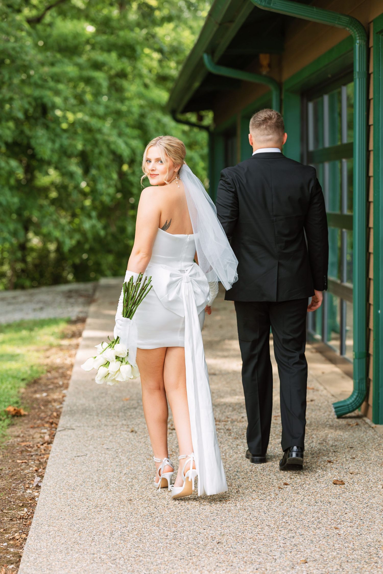Bride in white dress and groom in black suit walking together on a path with green pillars.