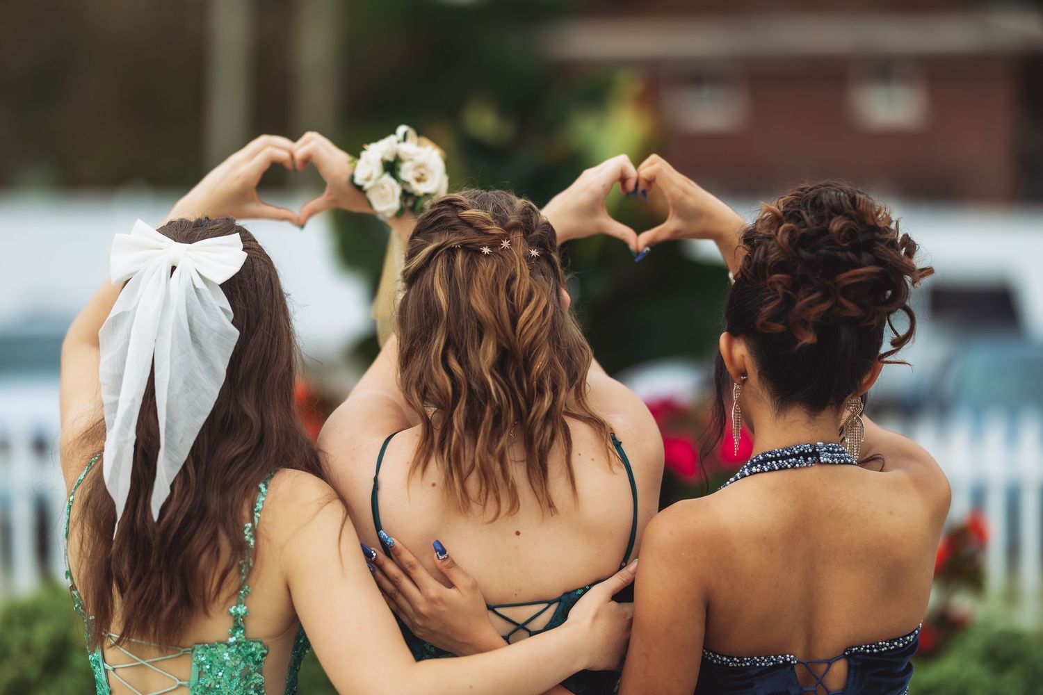 Three friends making heart shapes with their hands while wearing formal dresses at an outdoor event.