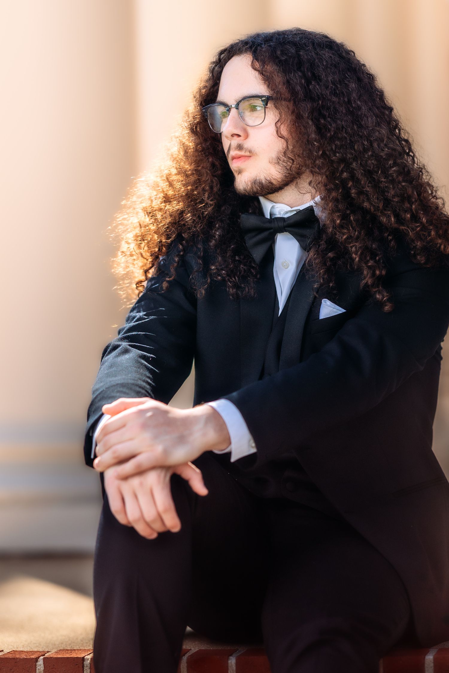 Elegant formal portrait of someone in a black tuxedo with bow tie, glasses and long curly hair in warm lighting.