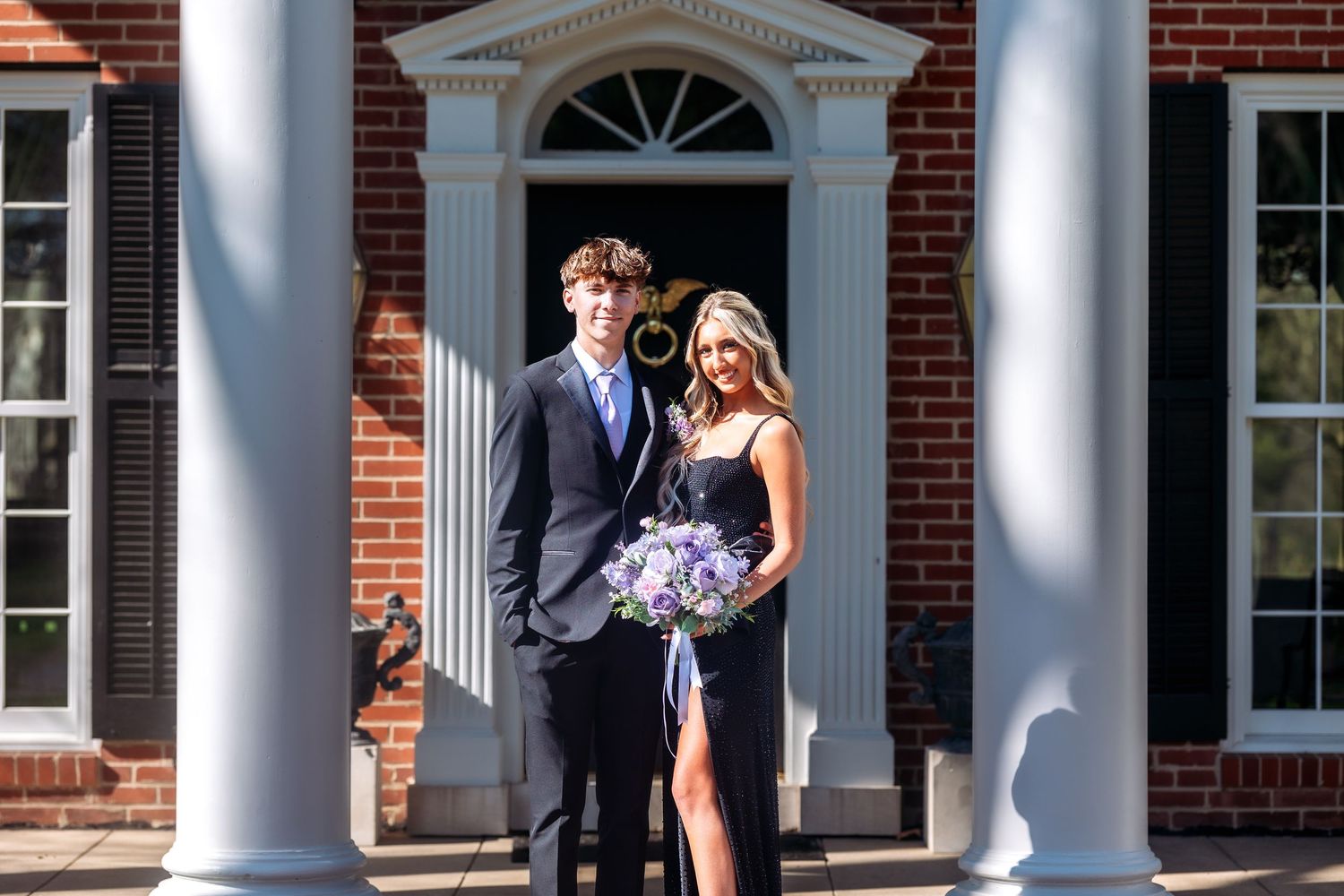 A couple stands elegantly between white columns at a formal entrance with a fanlight doorway of a brick building.