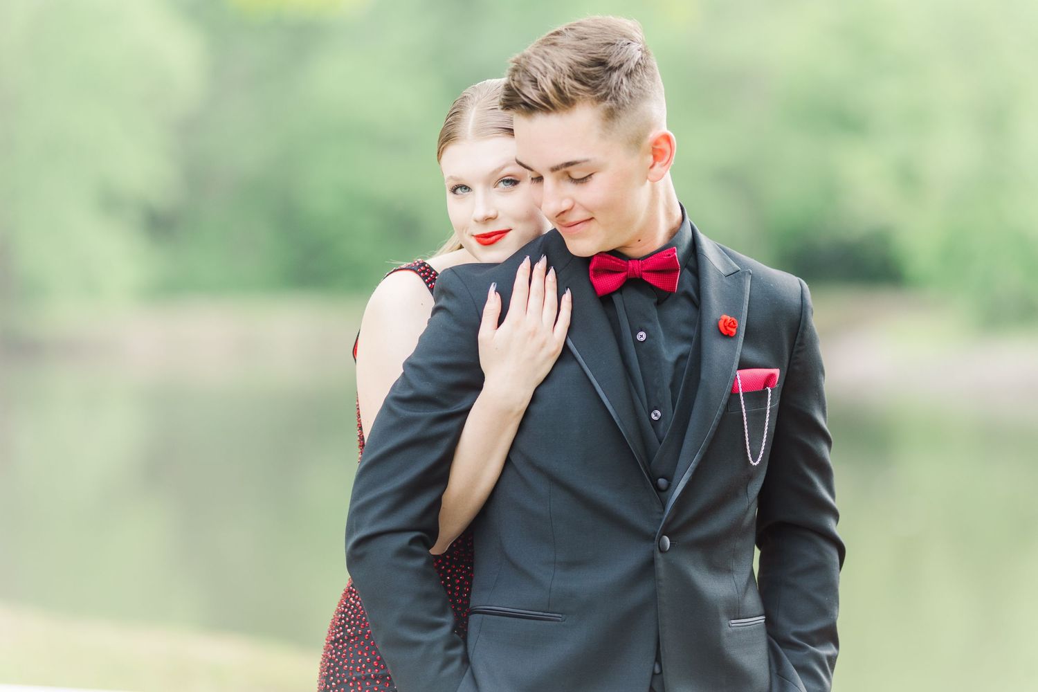 A formal couple shares an intimate moment outdoors wearing black suit and burgundy dress with red bowtie accent.