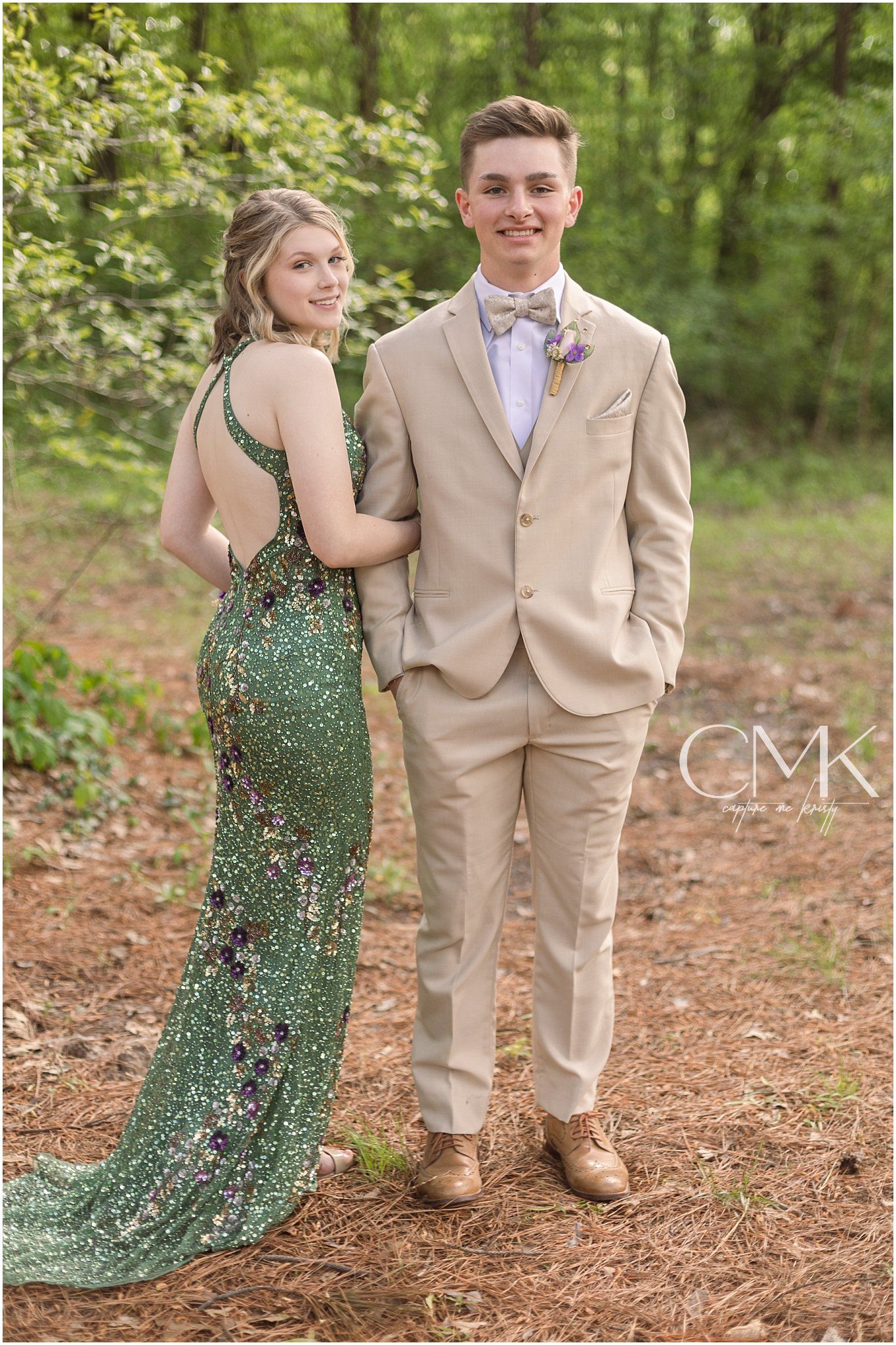 A couple poses for prom photos in a wooded setting, wearing a sparkly green gown and tan suit.