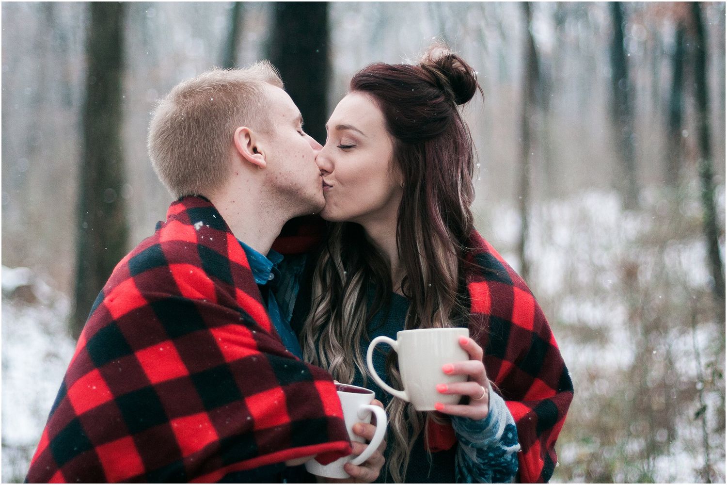 Cozy winter couple sharing coffee and kisses wrapped in a buffalo plaid blanket.