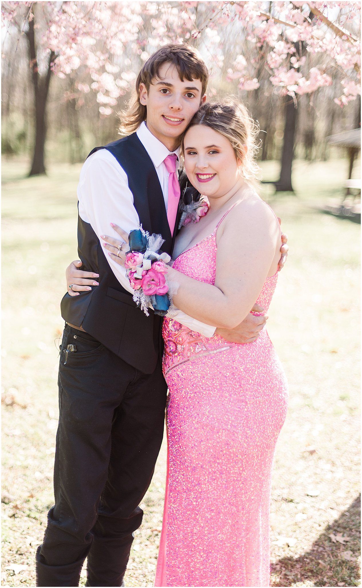 A couple poses in formal attire with a corsage under blooming spring trees.