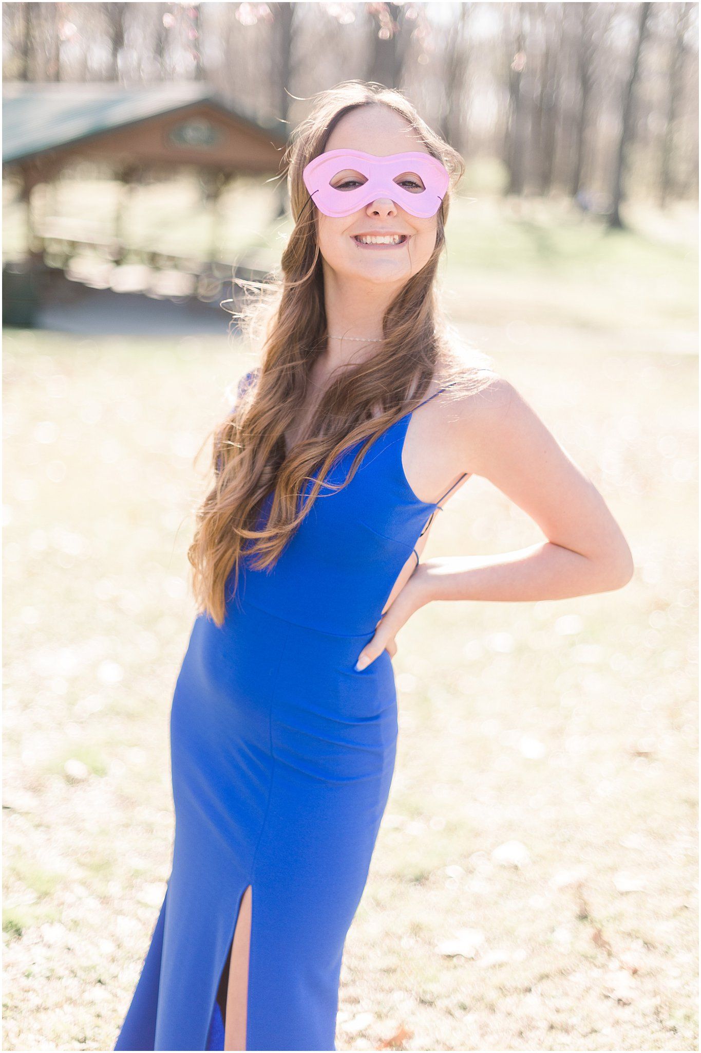 A person in a royal blue formal dress strikes a pose while wearing a pink masquerade mask outdoors.