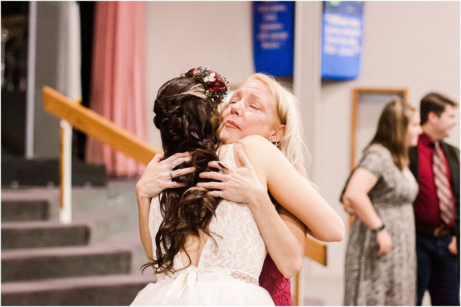Two people sharing an emotional embrace at a formal event with others in the background.