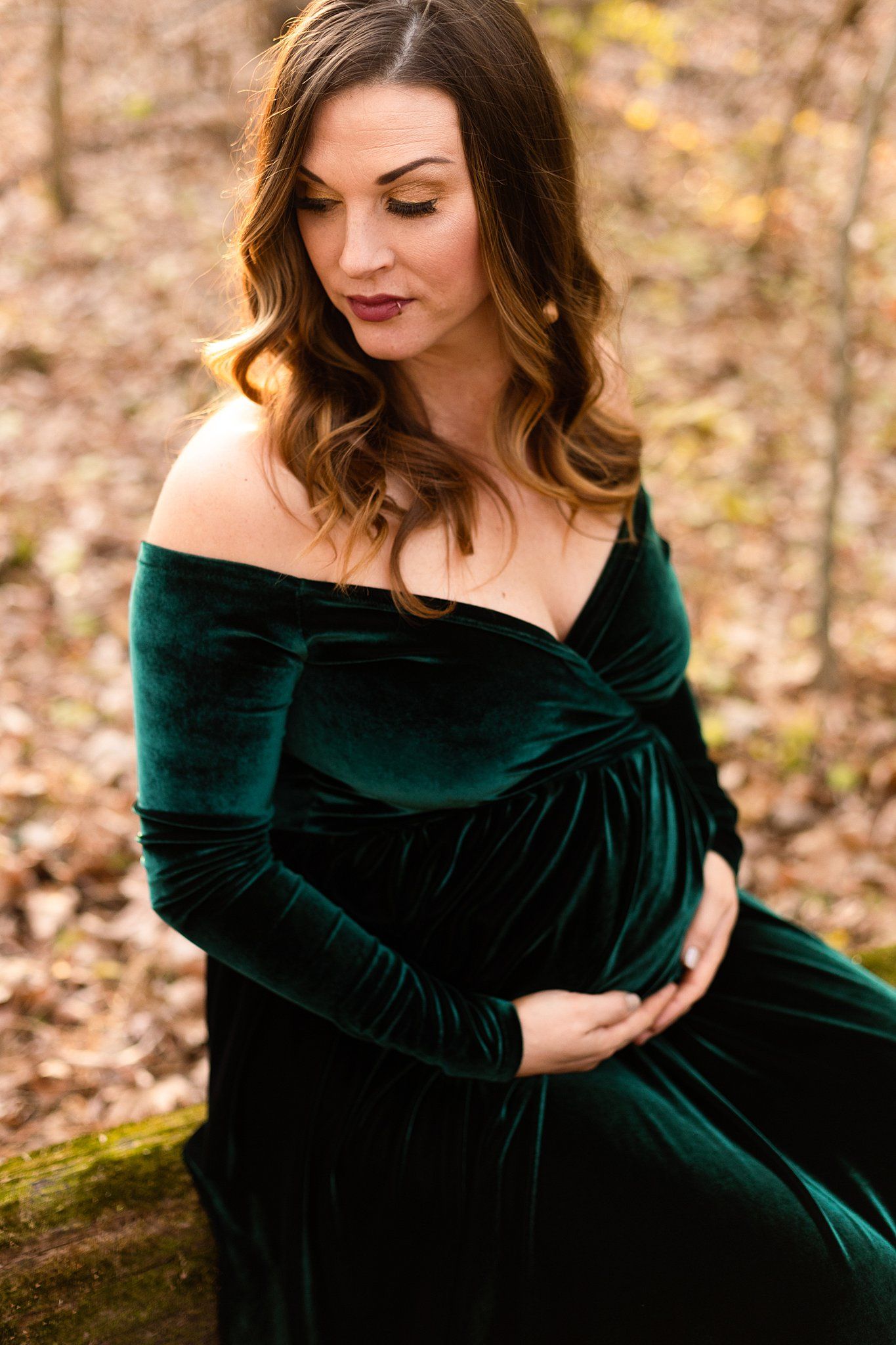 Elegant maternity portrait in emerald velvet off-shoulder gown against autumn backdrop.