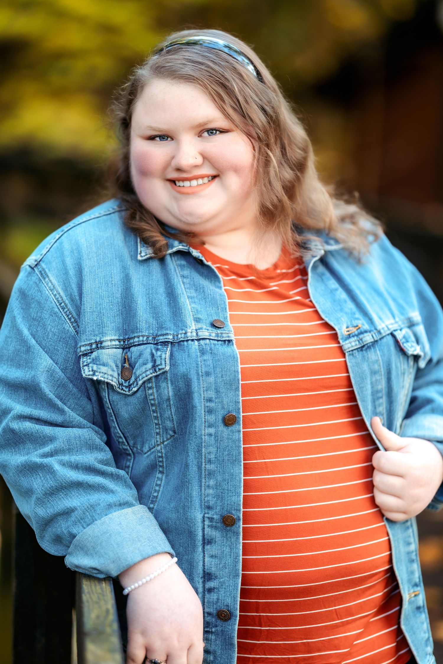 Person in denim jacket and orange striped shirt smiles during outdoor photoshoot.