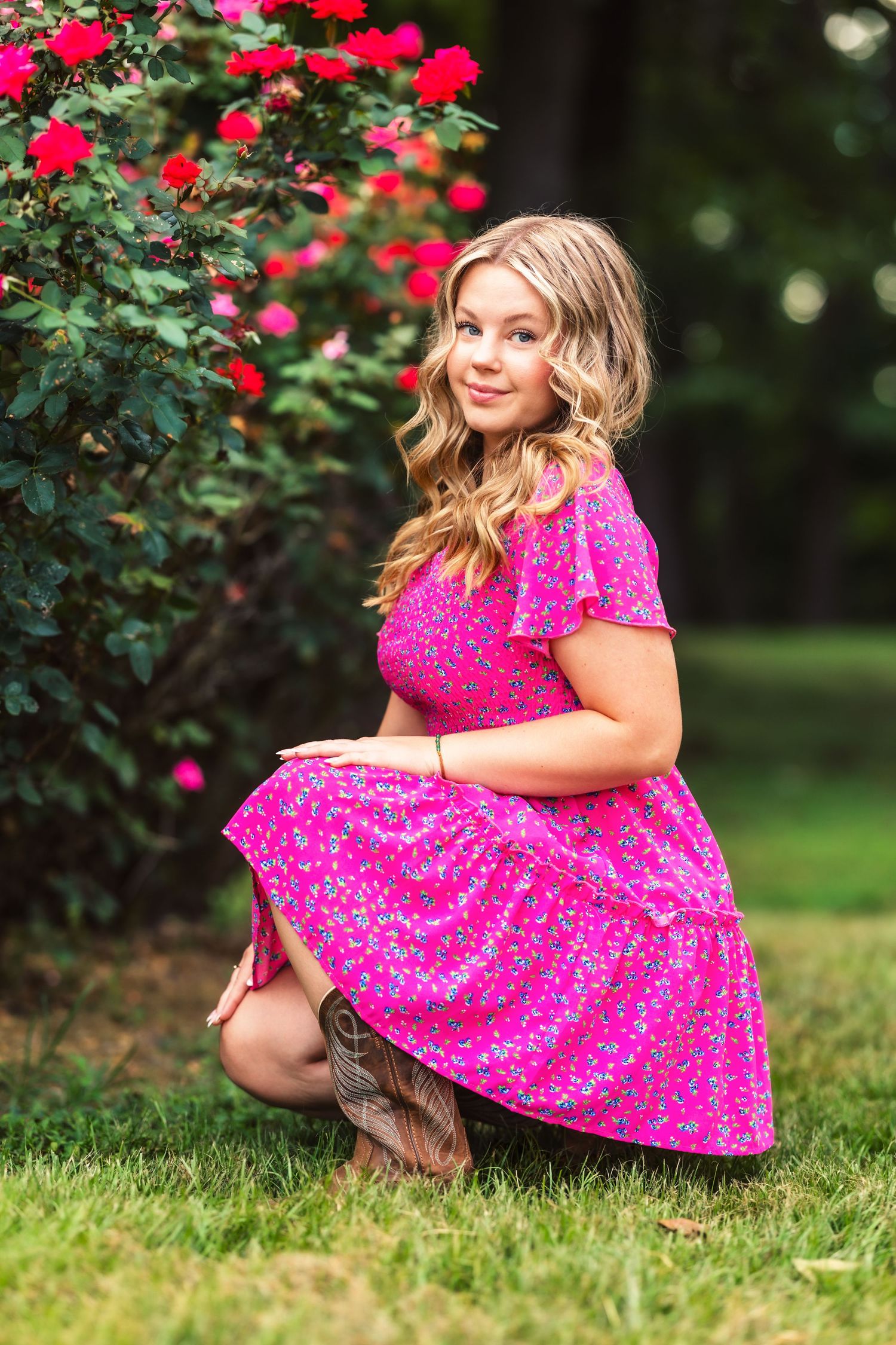Young girl in bright pink floral dress sits gracefully near blooming red roses in a garden setting.
