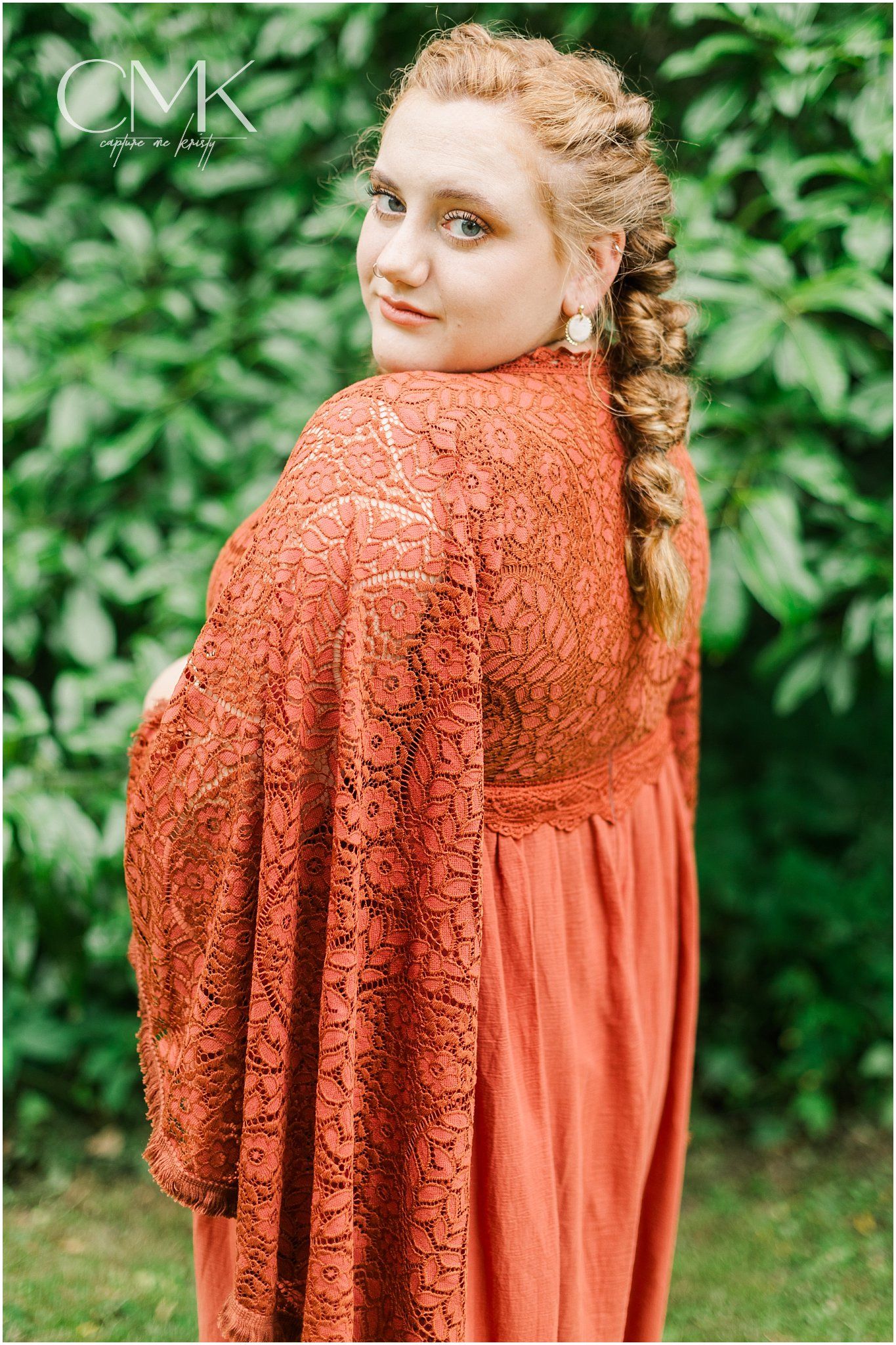 Side profile portrait in coral lace maternity dress with braided hair against green foliage.