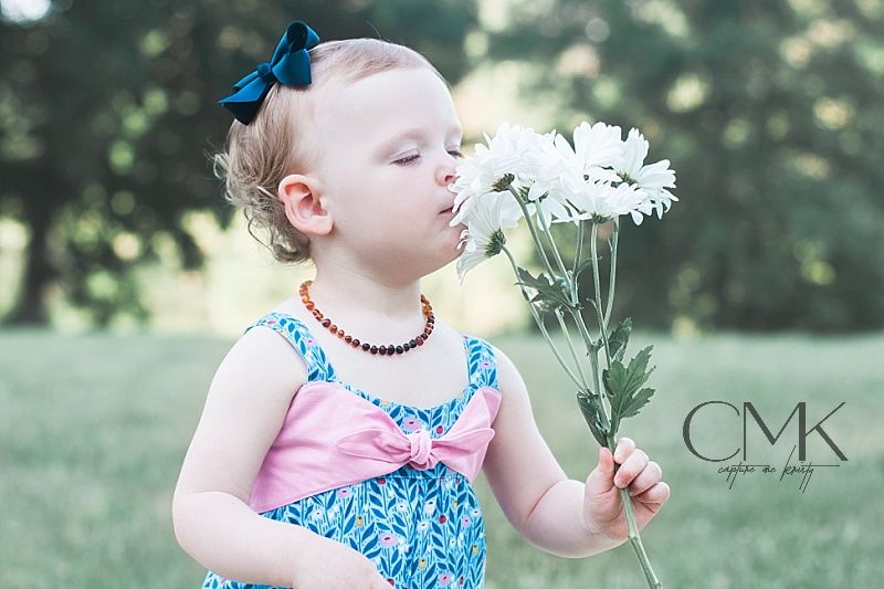 A small child in blue and pink dress smells white daisies in an outdoor setting.