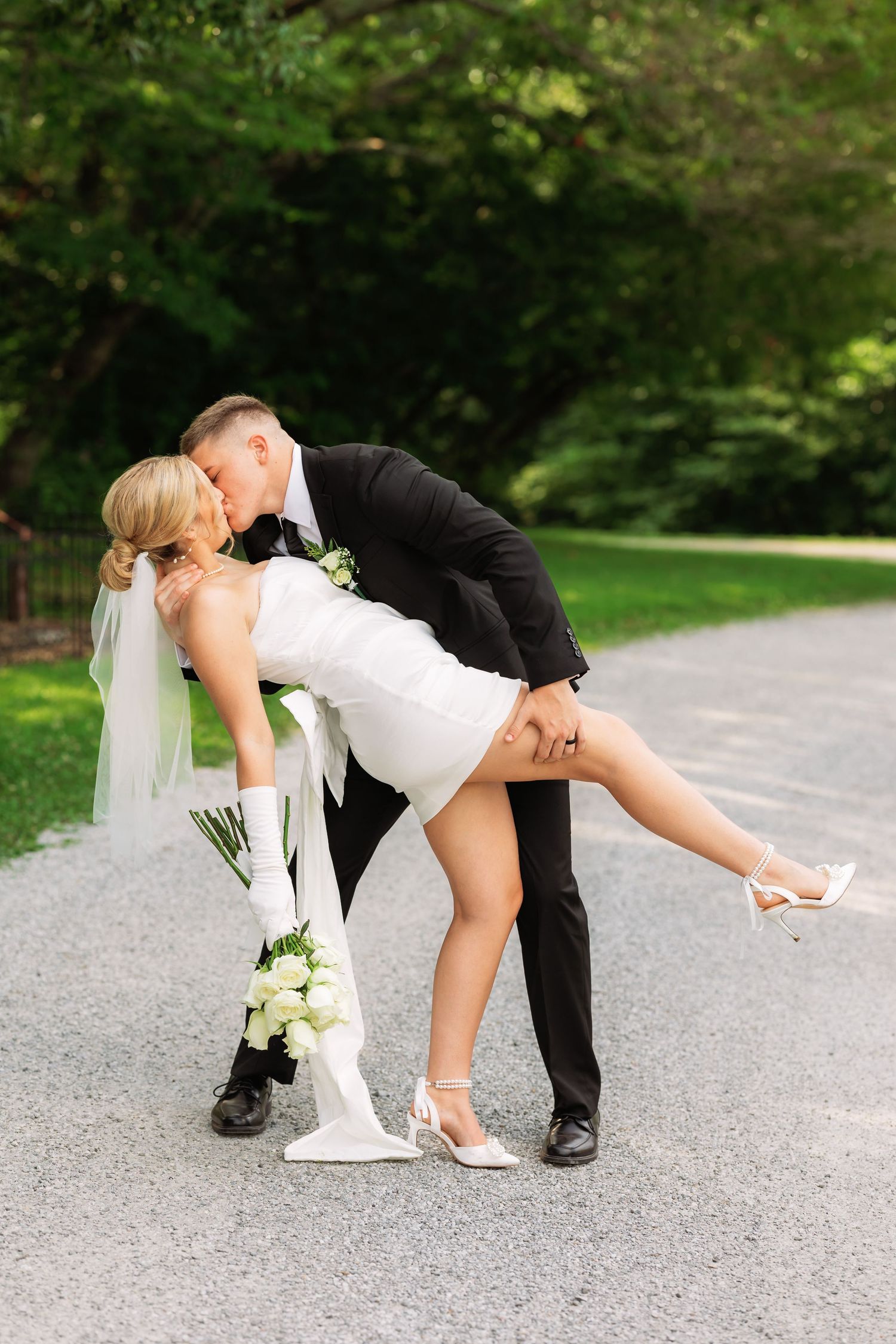 Playful wedding couple performs a romantic dip pose on a gravel pathway.