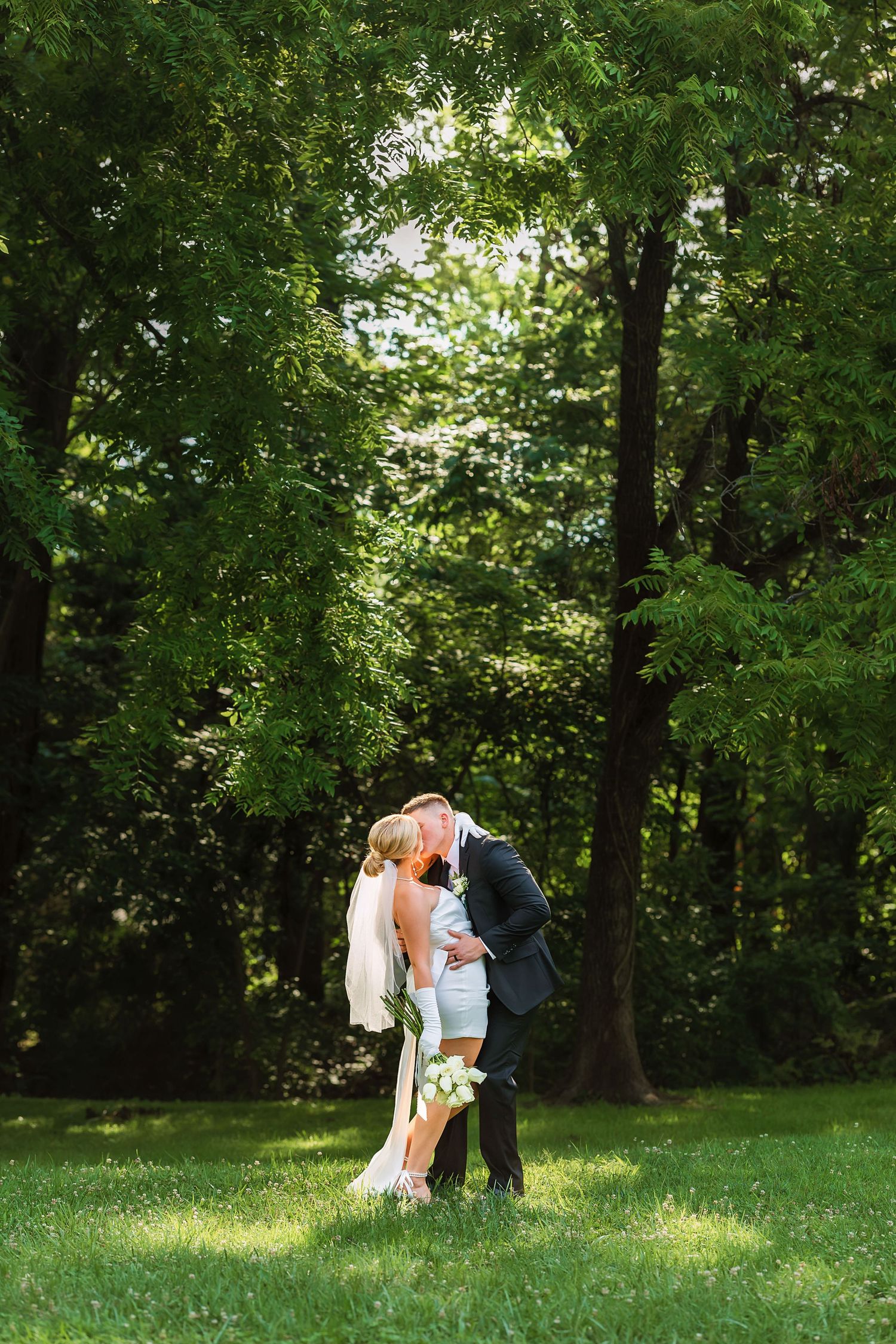 Newlyweds share intimate moments in a sunlit wooded garden setting.