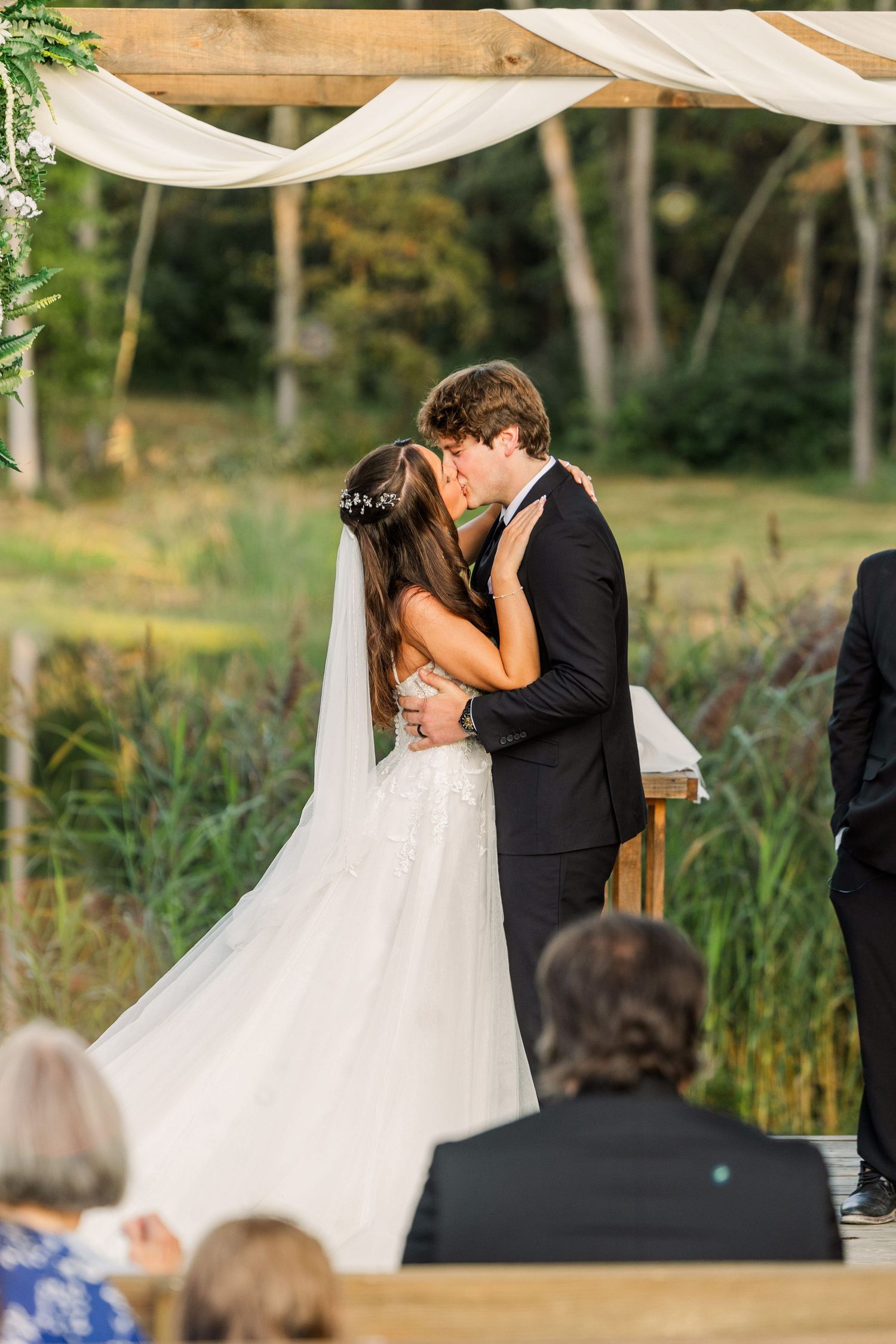 A romantic first kiss during an outdoor wedding ceremony under a draped canopy with guests watching.