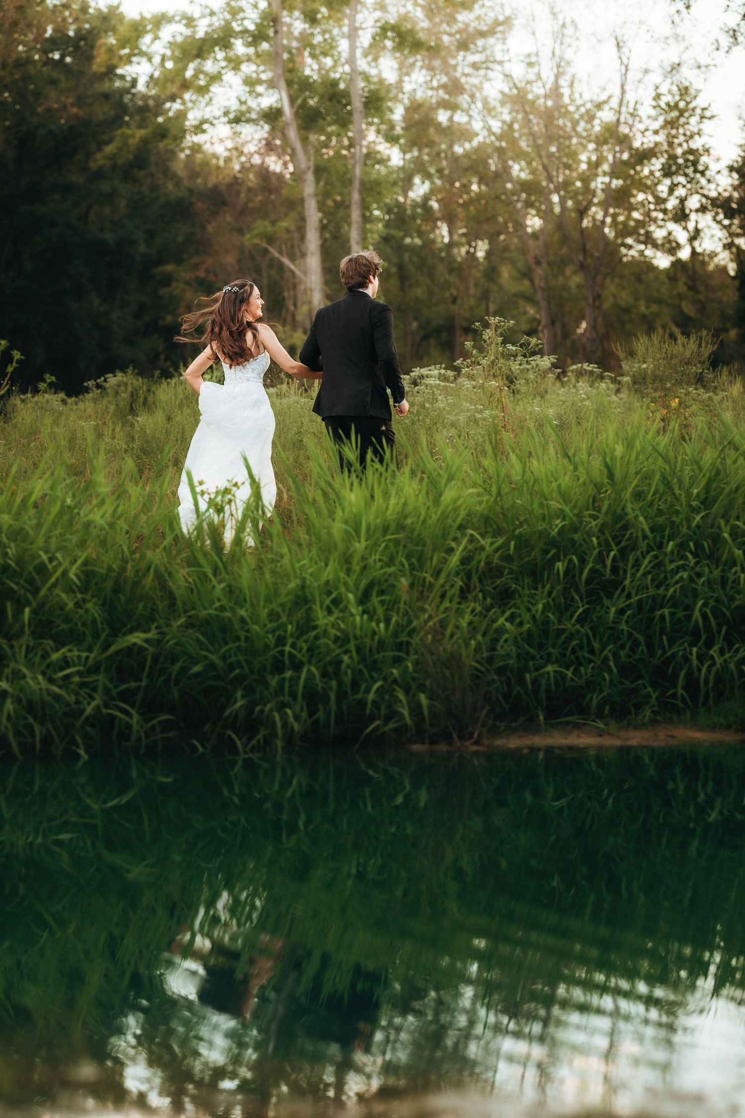 A romantic wedding couple walking through tall grass beside a reflective pond at sunset.