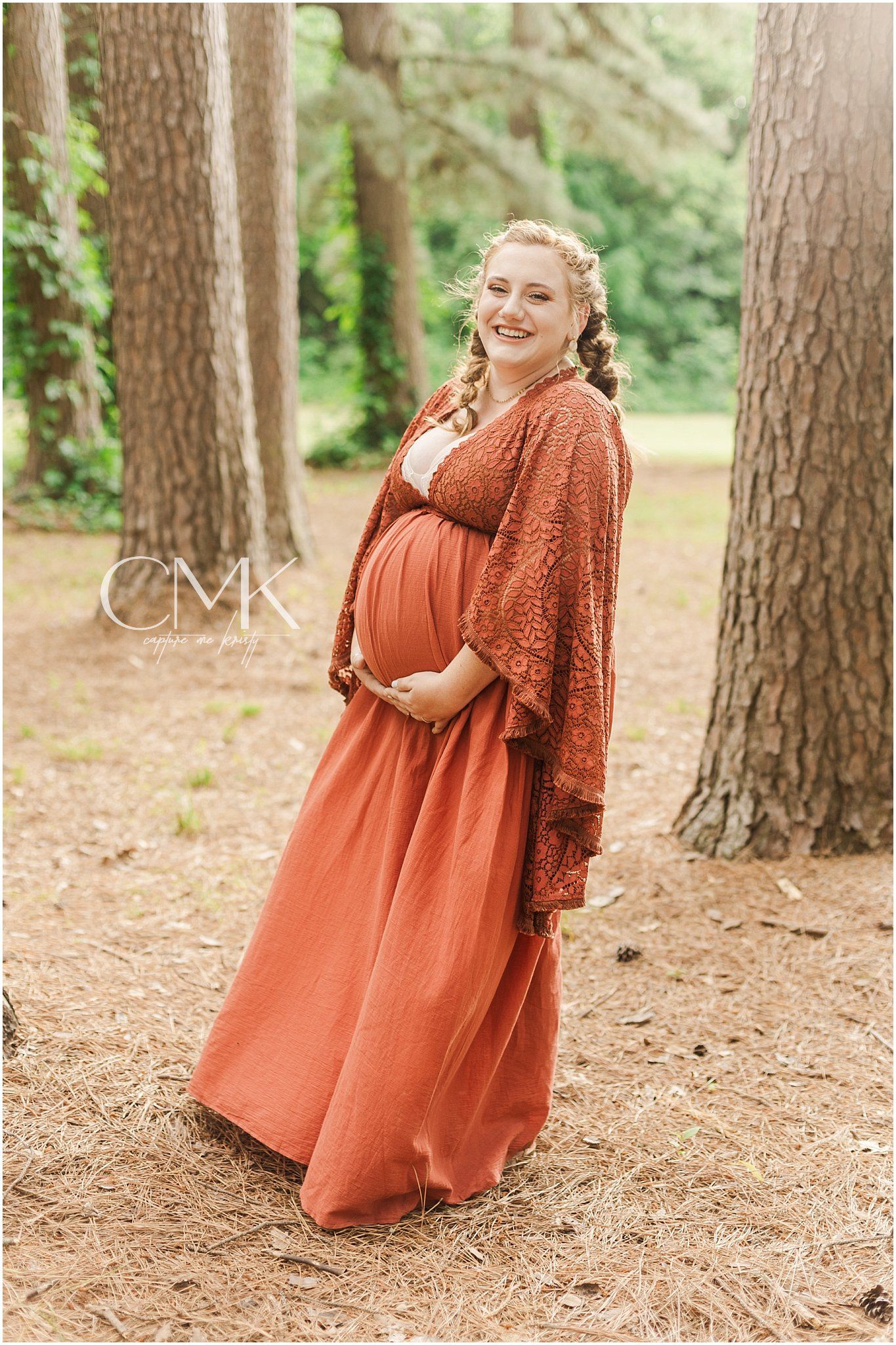Elegant maternity photo showing a flowing rust dress and lace overlay in a pine forest.