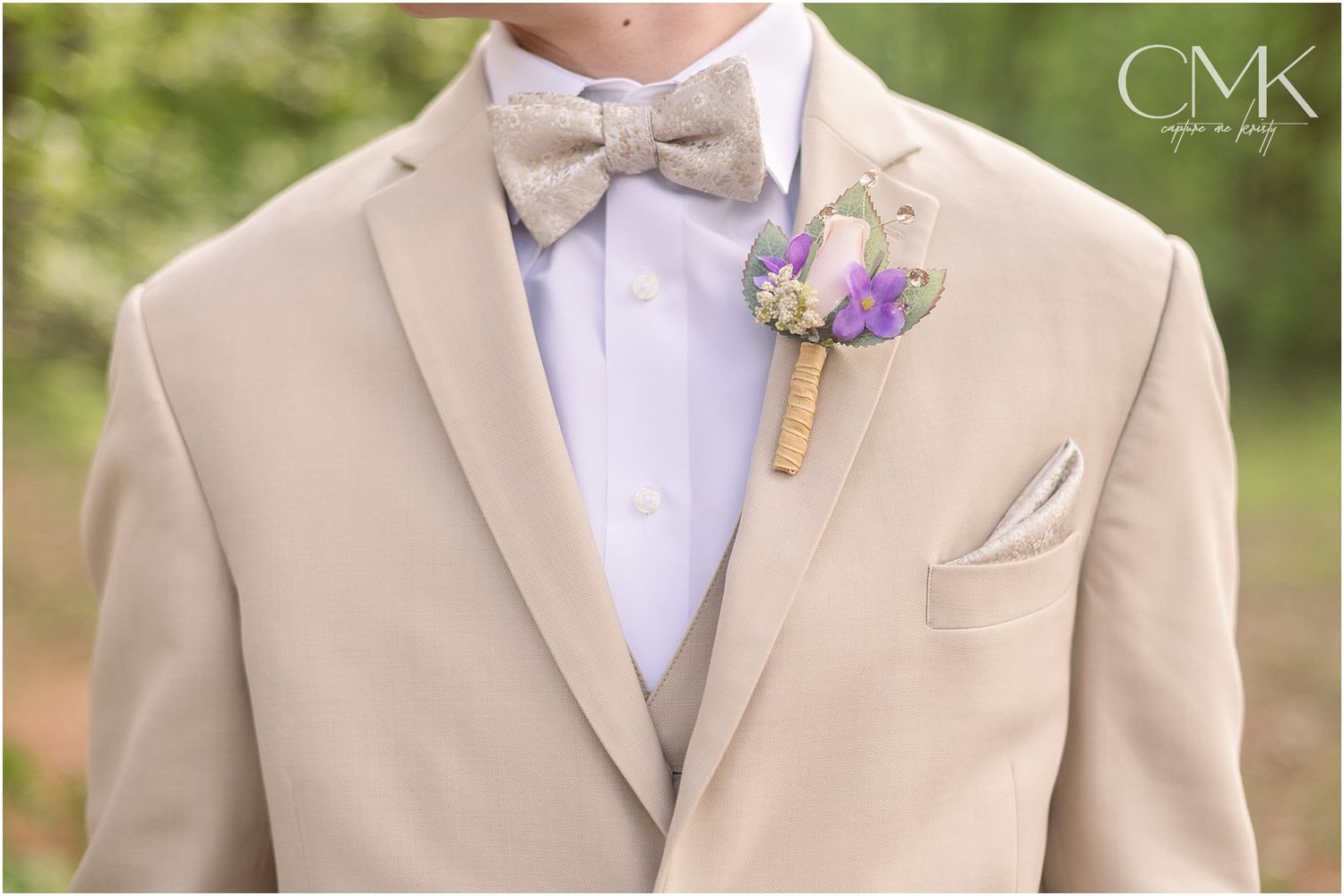 Close-up details of a beige suit jacket with purple boutonniere and white bow tie.