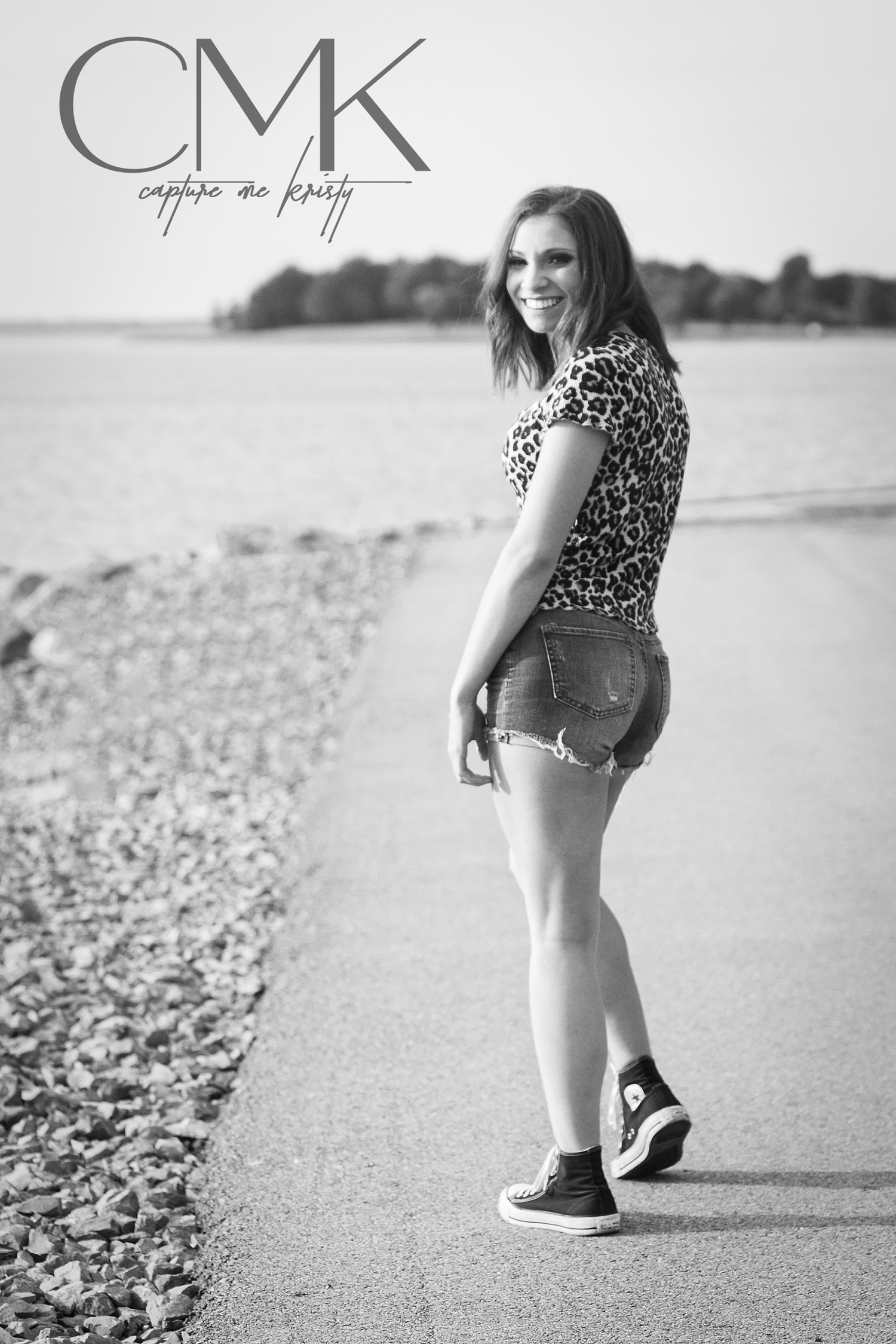 Black and white portrait showing casual pose in leopard print top and shorts along rocky waterfront path.
